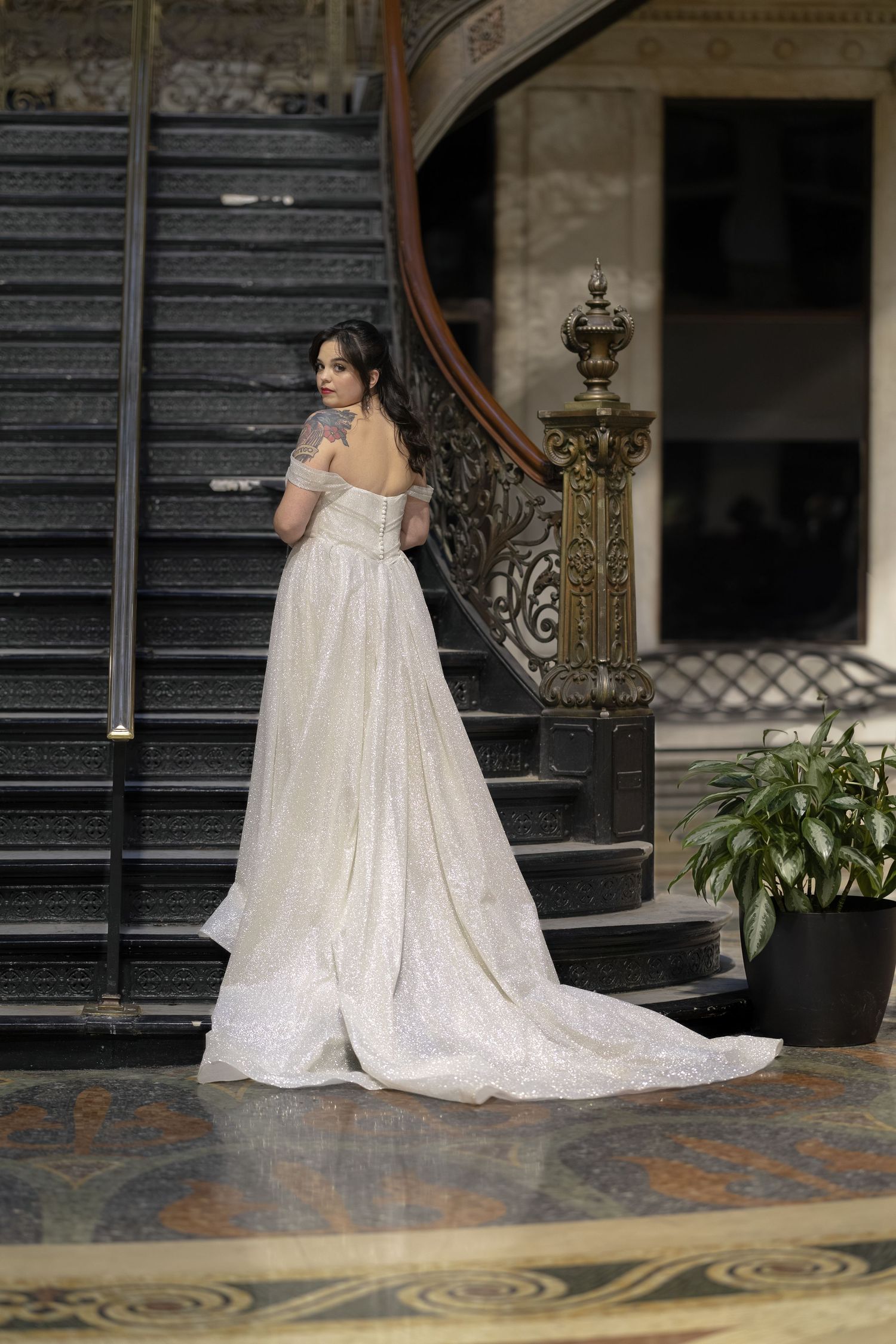 A bride in a flowing white sequined wedding gown stands elegantly on ornate vintage stairs with decorative railings.