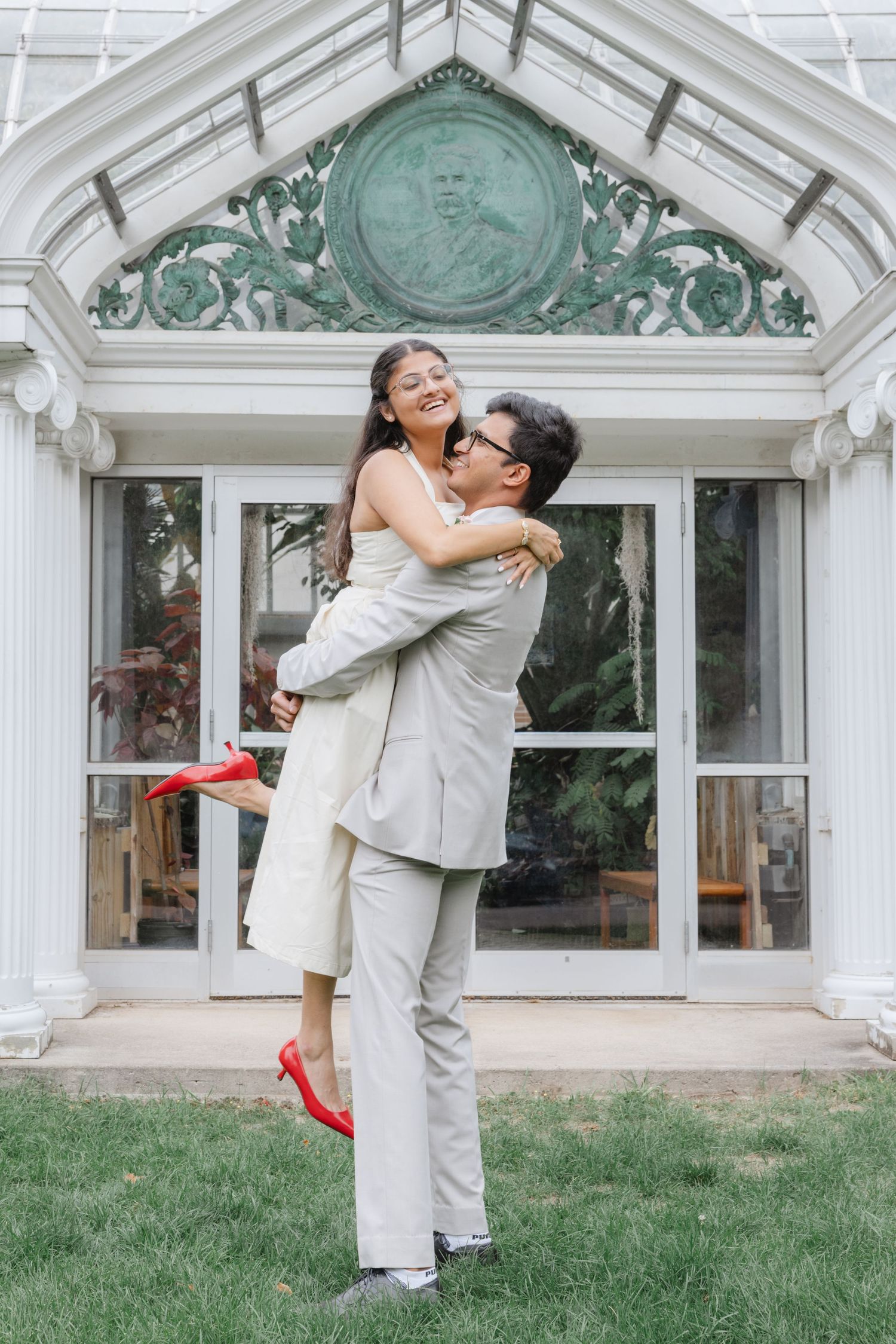A couple in wedding attire share a romantic embrace on a lawn in front of a white conservatory building.