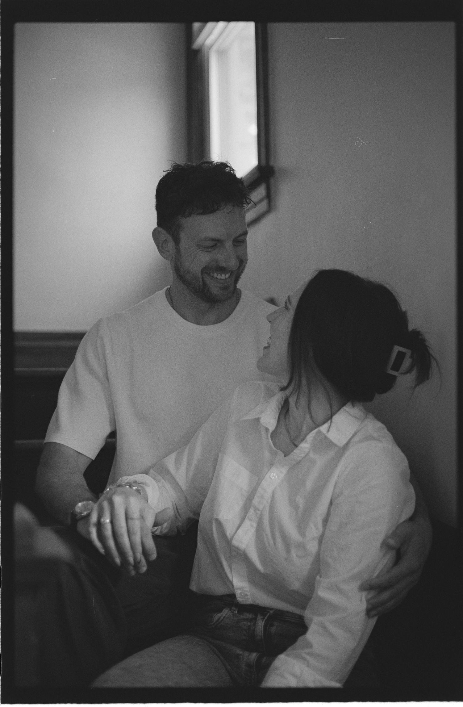 Black and white photo of a smiling man and woman sitting together, sharing a tender moment indoors.