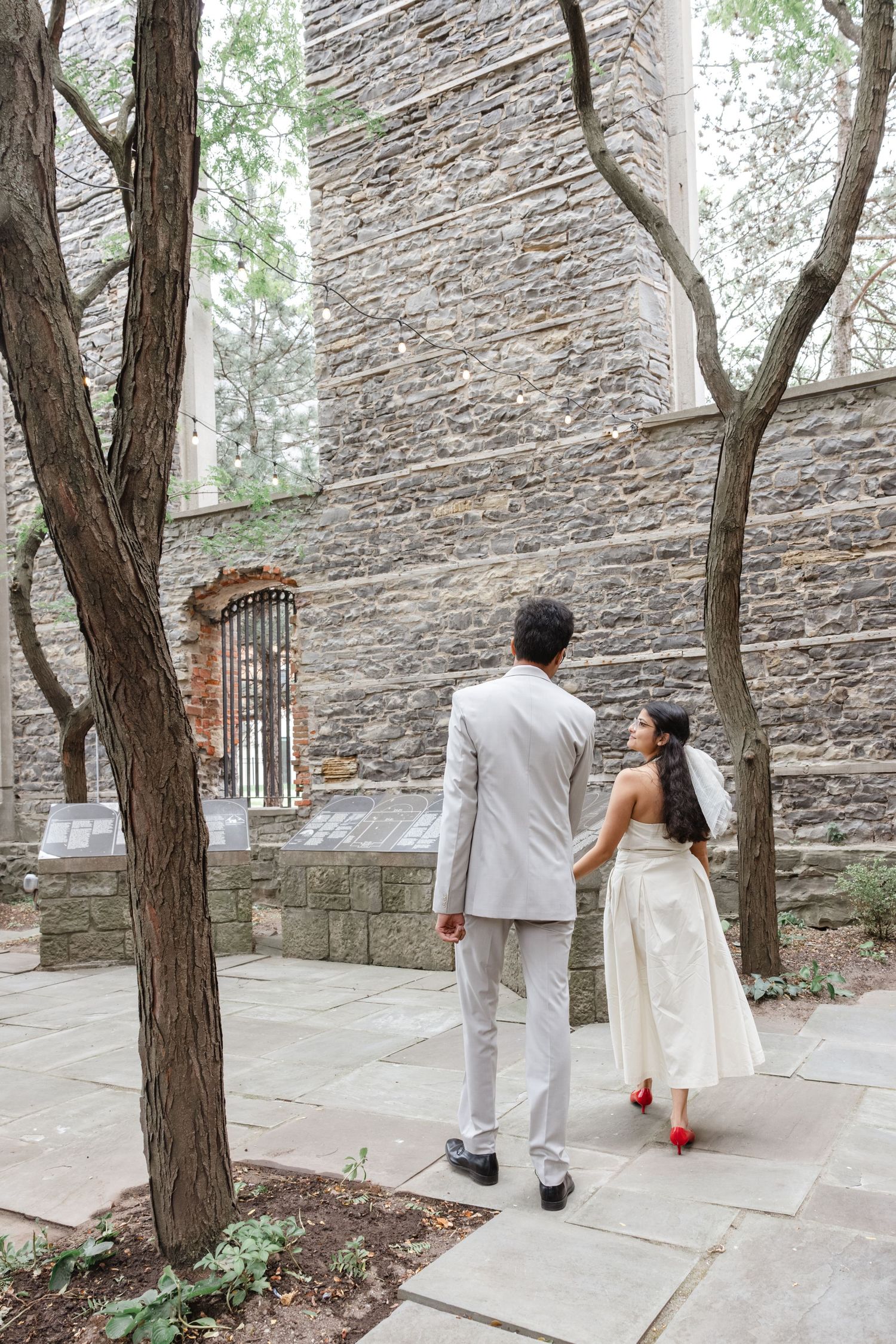 A couple walks along a stone path beside a historic building with trees framing the scene.  St. Josephs park wedding photographer