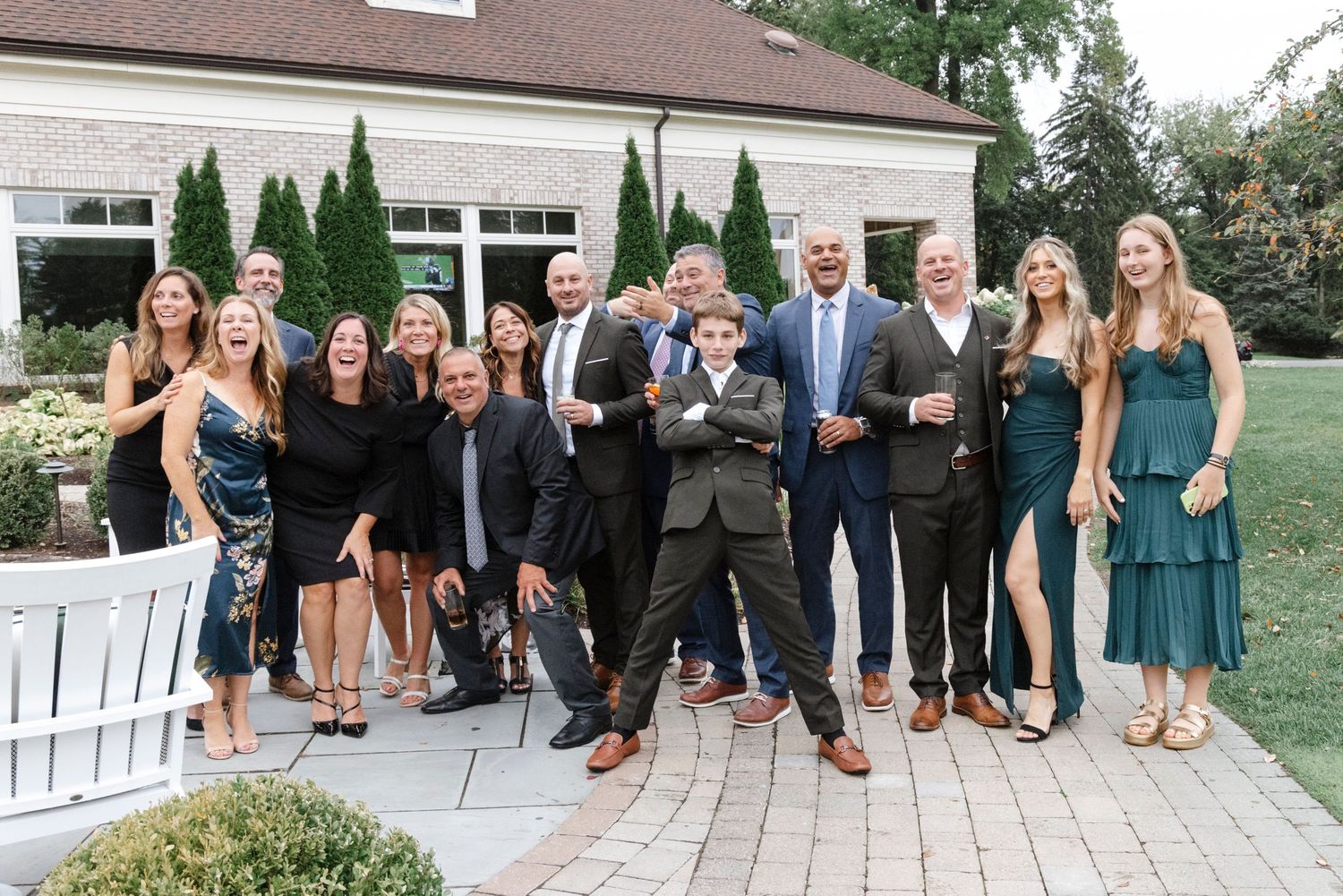 Wedding party members in formal attire gather outdoors on a lawn with evergreen trees in the background. Rochester NY documentary wedding ph
