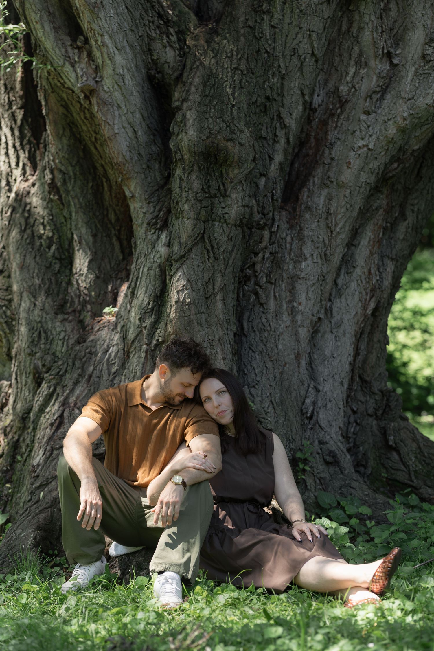 Couple sitting together against large ancient tree trunk in lush green park, man in brown shirt embracing woman in dark dress.