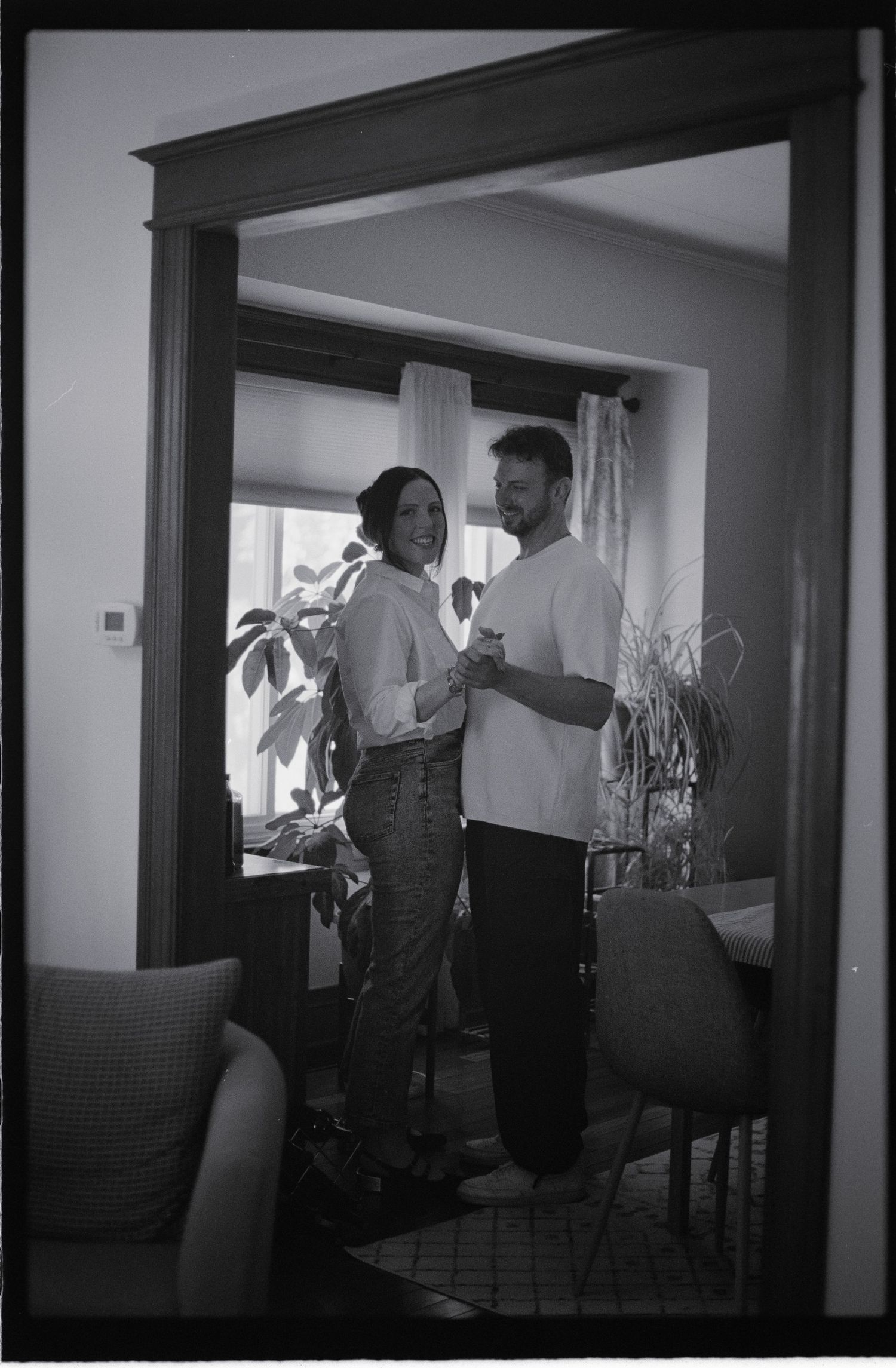 Black and white photo of a couple dancing and laughing together in a cozy home doorway surrounded by plants.