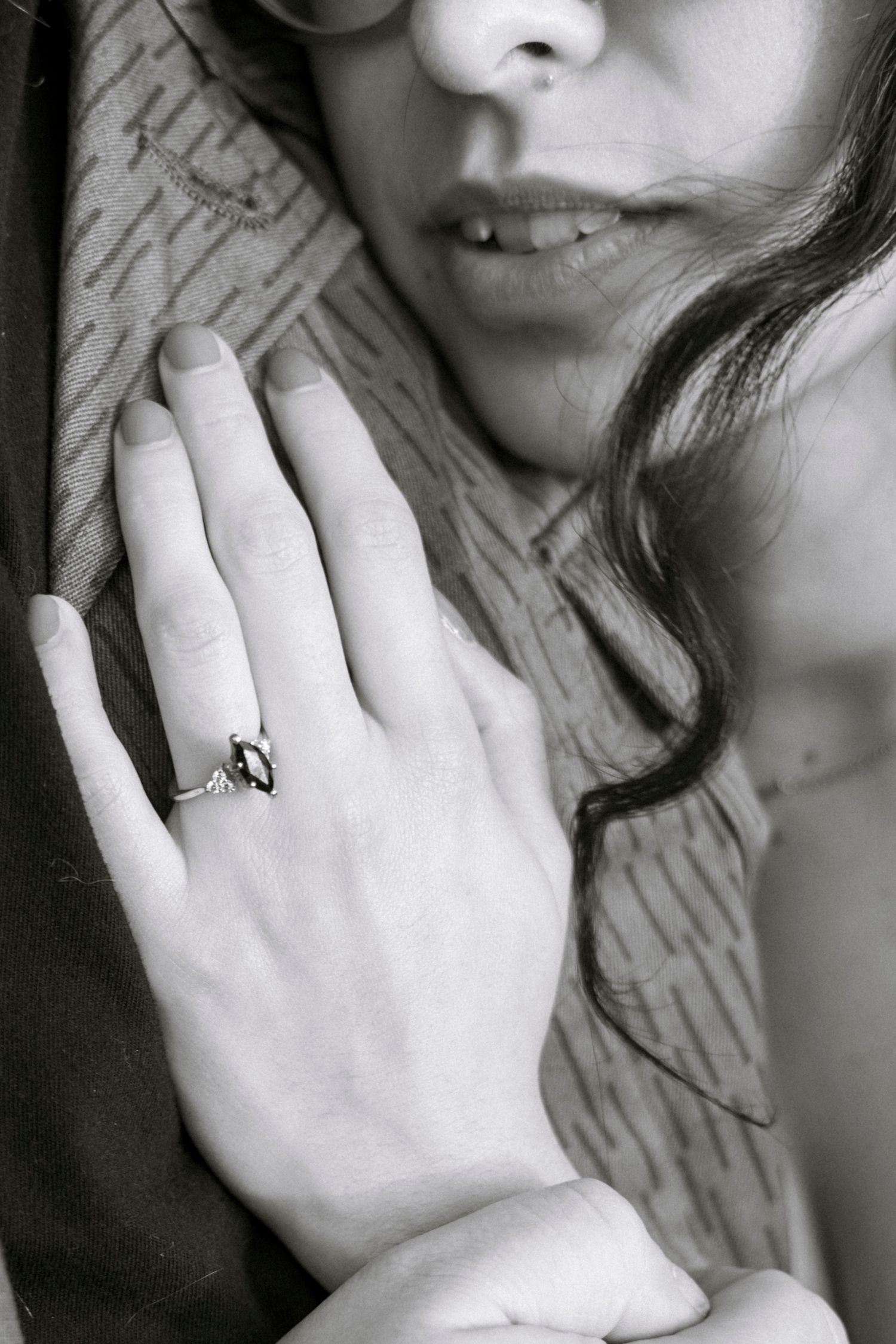Black and white close-up of a hand wearing an engagement ring with dark wavy hair and lips in soft focus.