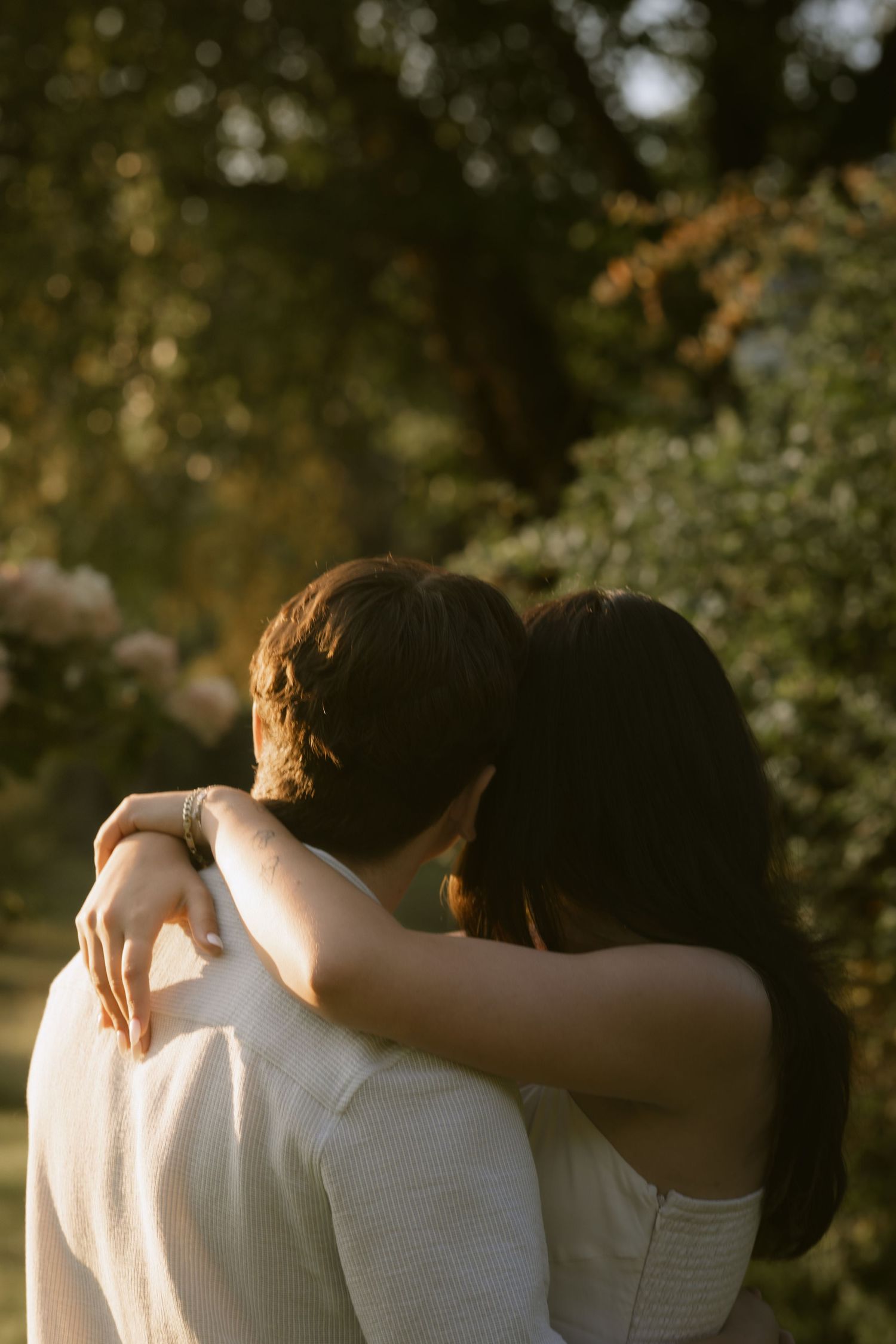 Two people embracing in white clothing against a blurred natural background during golden hour. Rochester NY authentic engagement photograph