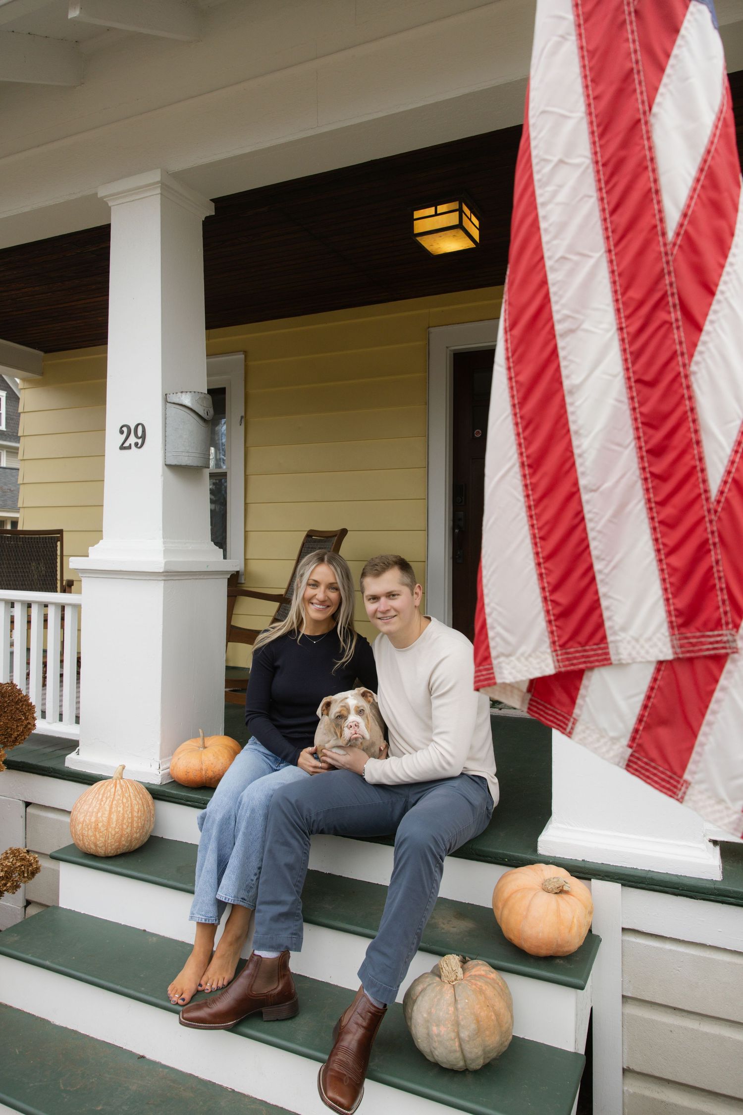 A couple sits with their dog on porch steps with American flag and pumpkin decorations. Rochester NY home engagement photography