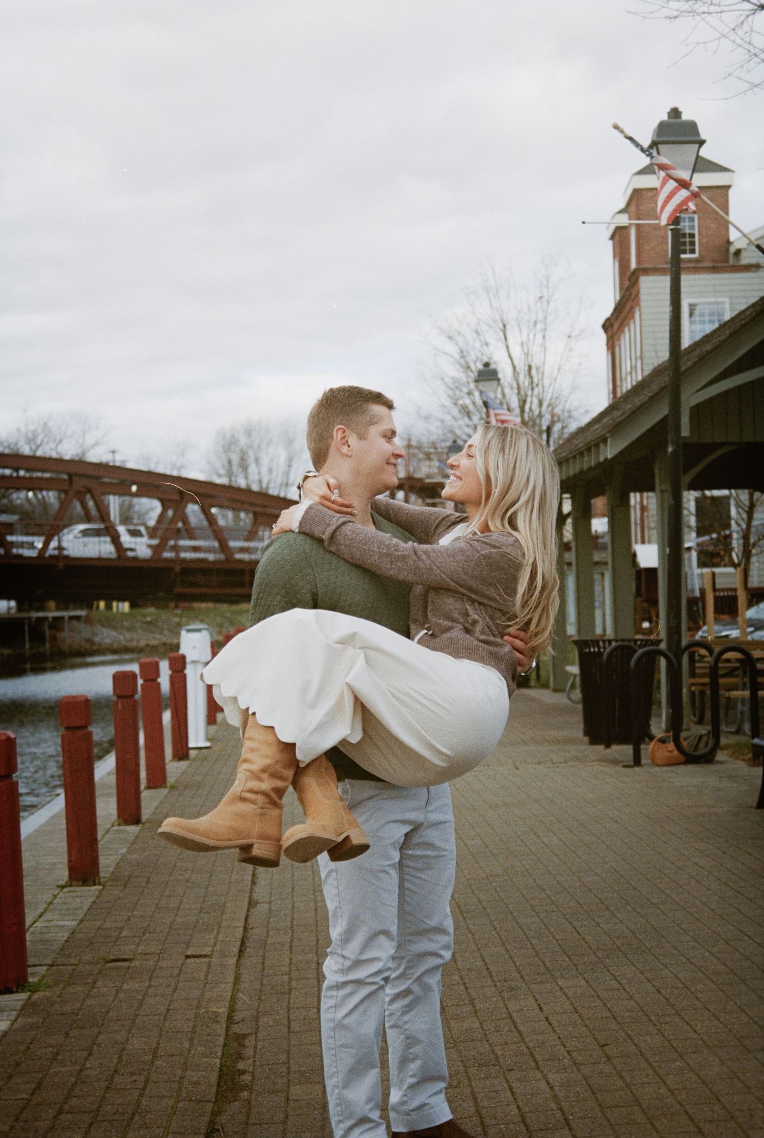 A romantic couple in casual attire share an intimate dip pose on a brick-paved boardwalk near vintage buildings.