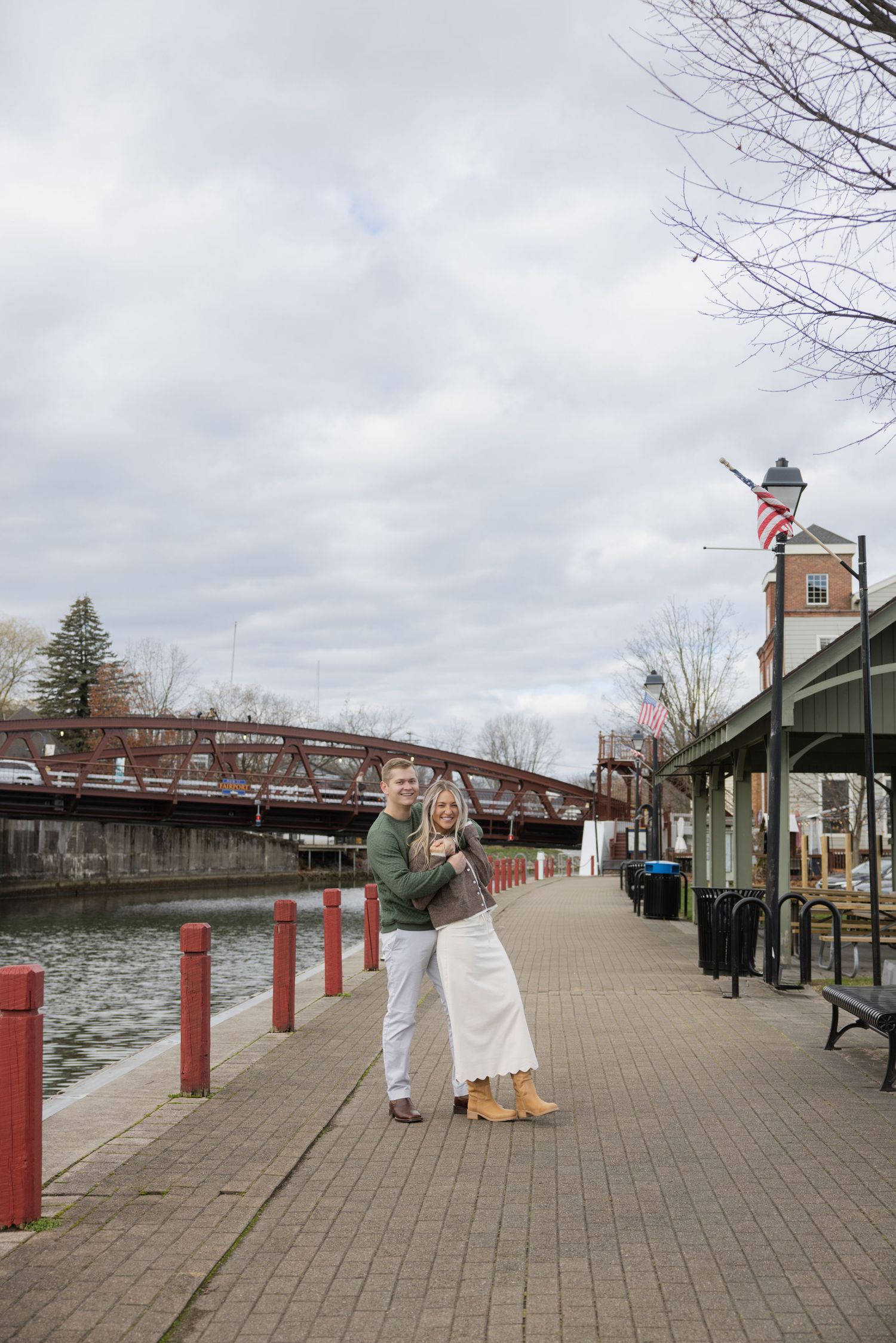 A couple stands together on a riverside walkway with a historic iron bridge and American flag in the background. Film wedding photographer R