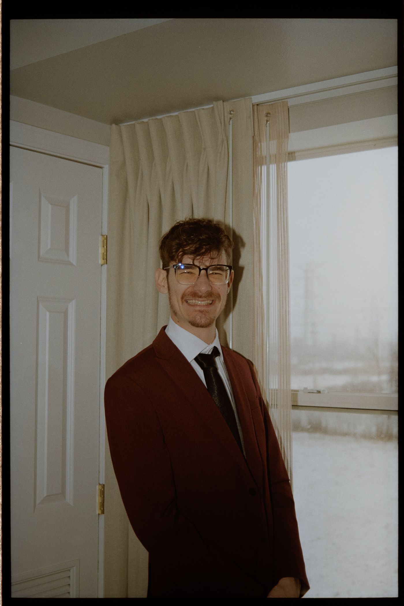A person wearing glasses, a burgundy suit jacket and black tie stands smiling by a white door in natural lighting.