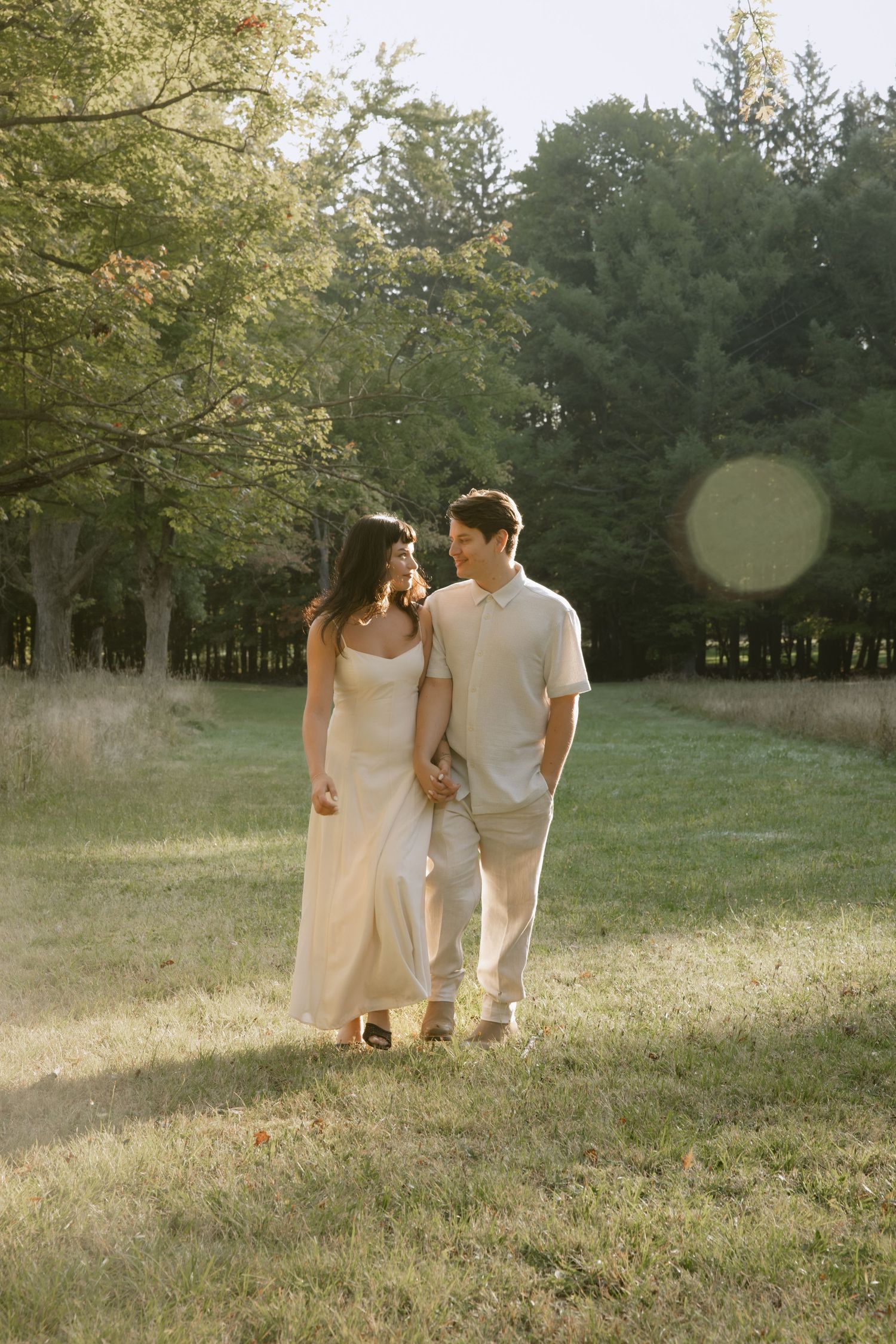 A couple walks hand in hand through a sun-drenched field with evergreen trees in the background.  Rochester NY film engagement photography