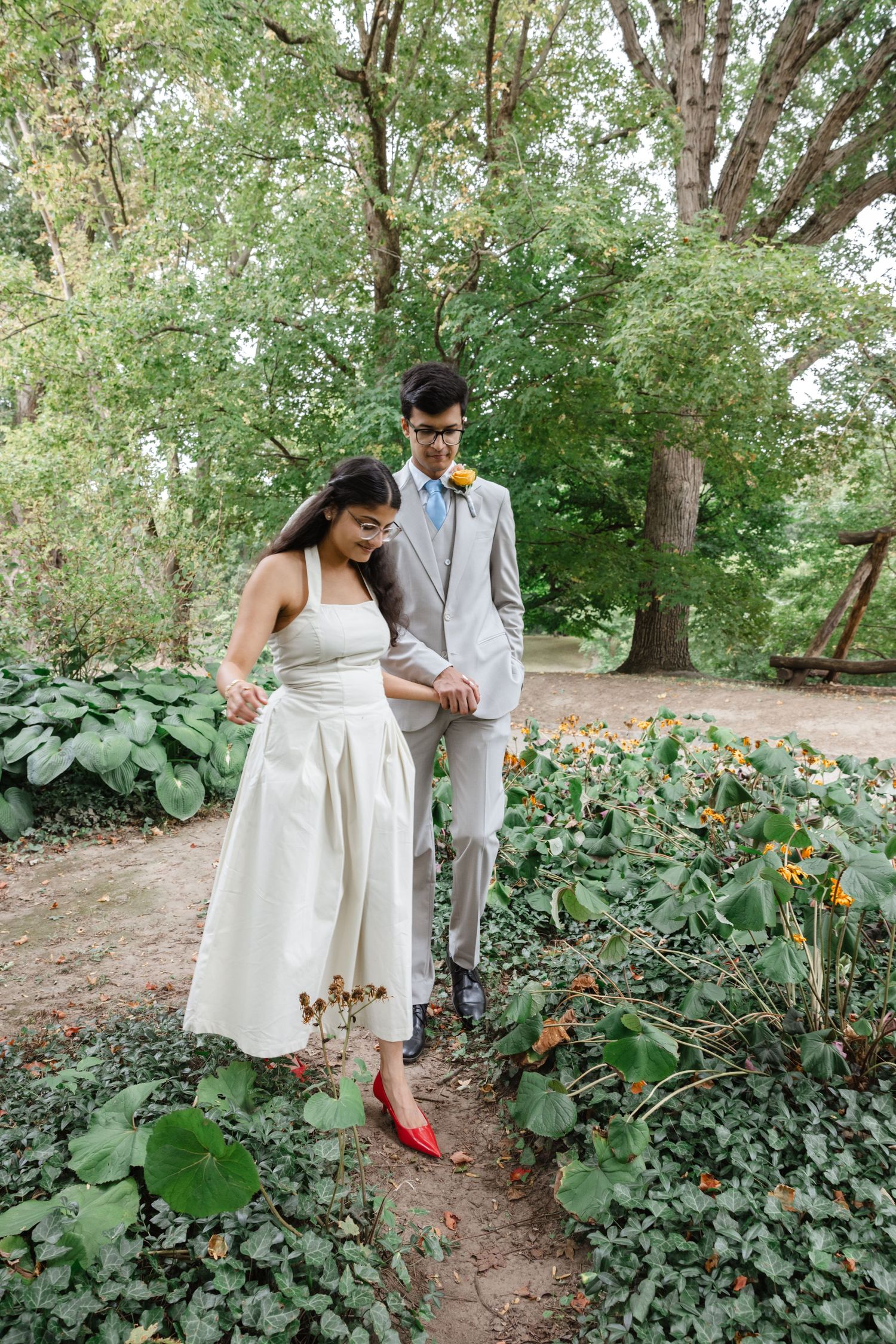 A couple strolls through a garden path with red flowers and lush greenery.  Rochester conservatory wedding photographer