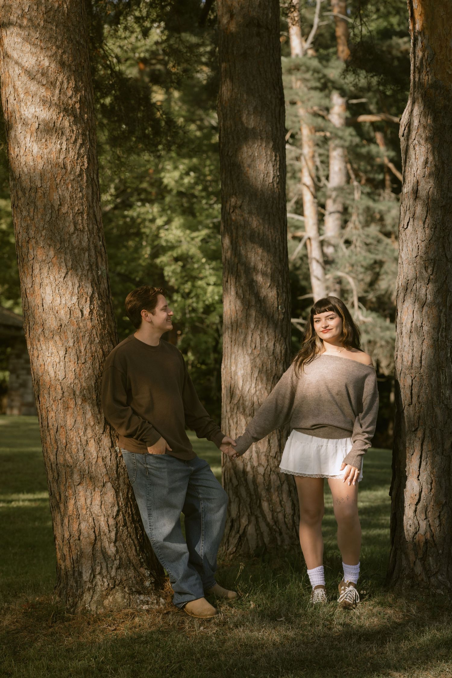 A casual couple poses against large tree trunks in a wooded setting.  Rochester NY film engagement photography