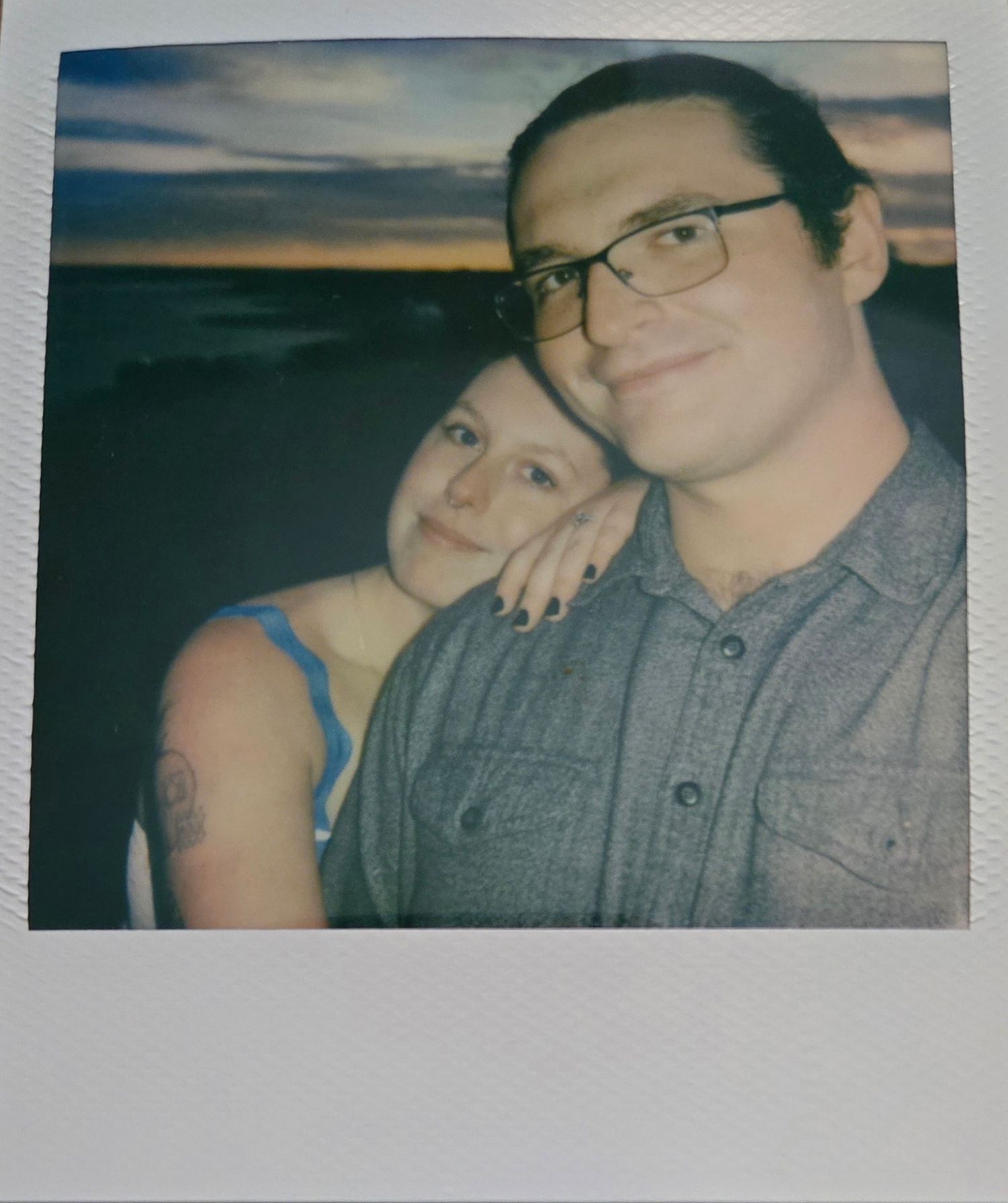 Couple posing for a Polaroid photo at sunset, man wearing glasses and gray shirt, woman in blue top with tattoo.