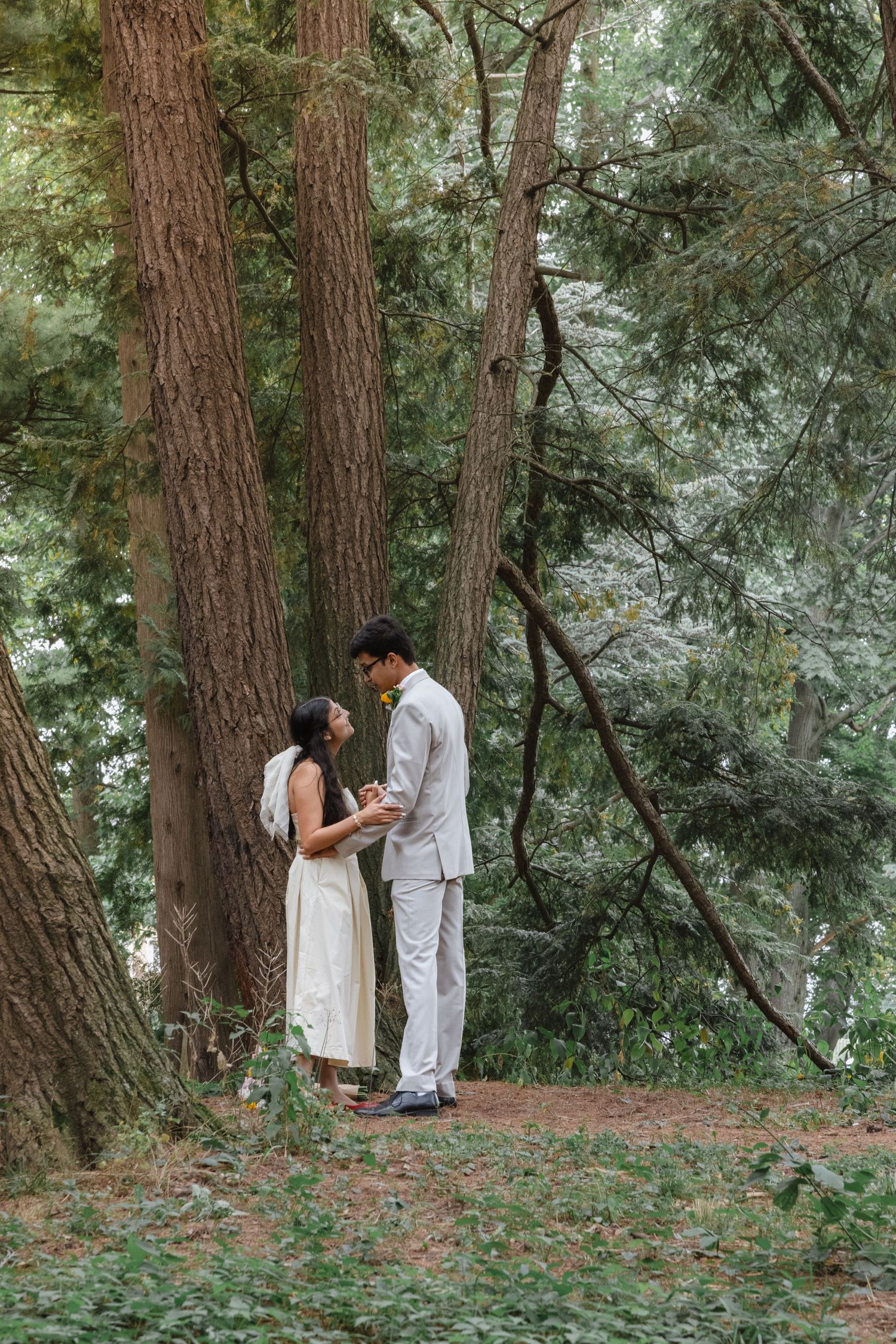 A couple embracing in a lush, forested environment with tall trees and greenery surrounding them.
