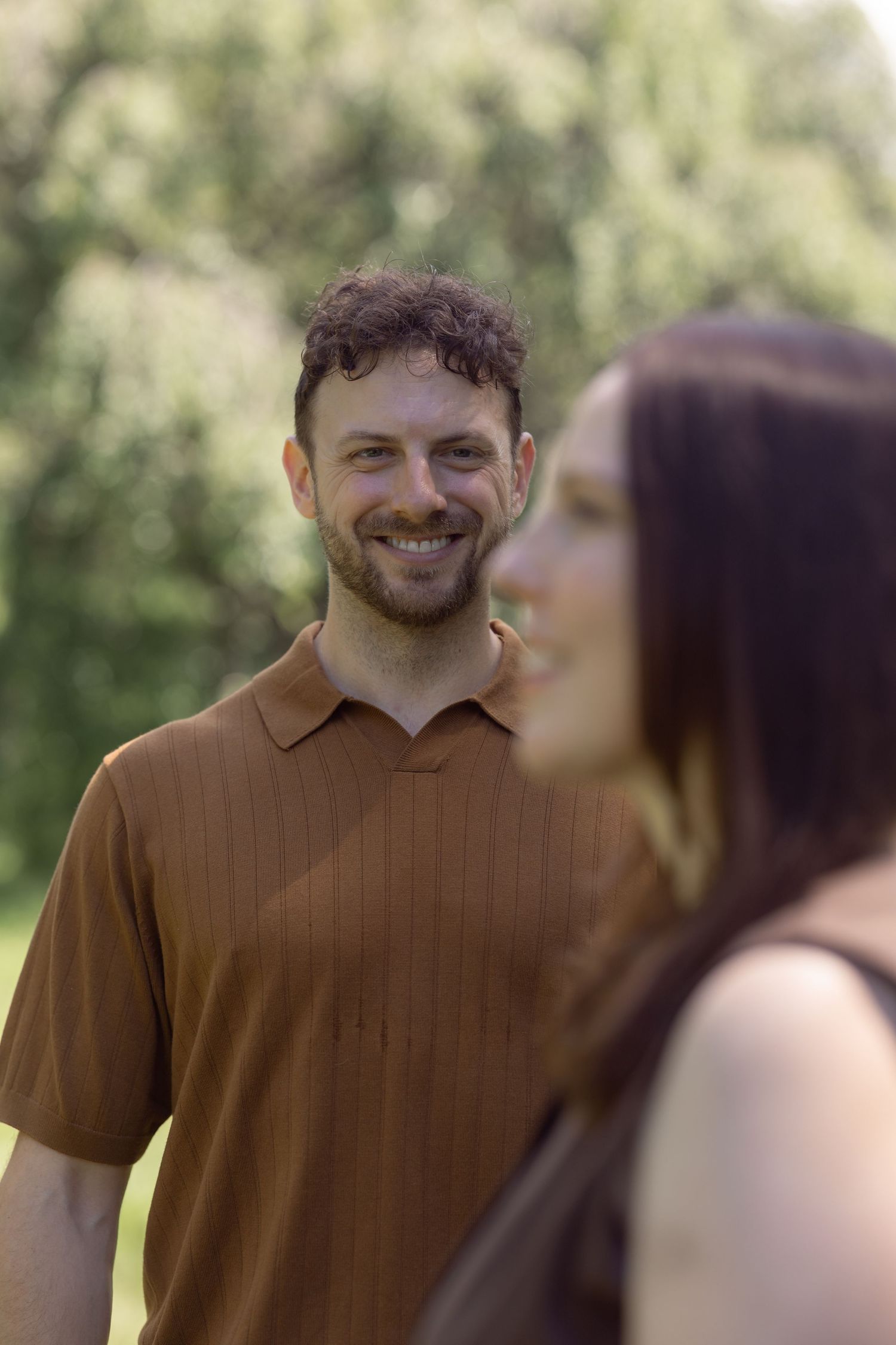 Smiling bearded man in brown polo shirt standing outdoors in park with blurred woman in foreground.