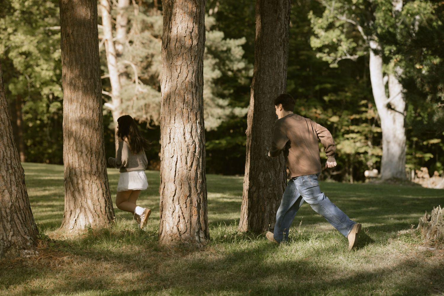 A couple playfully chases each other between tall trees in a sunlit park.  Rochester NY film engagement photography