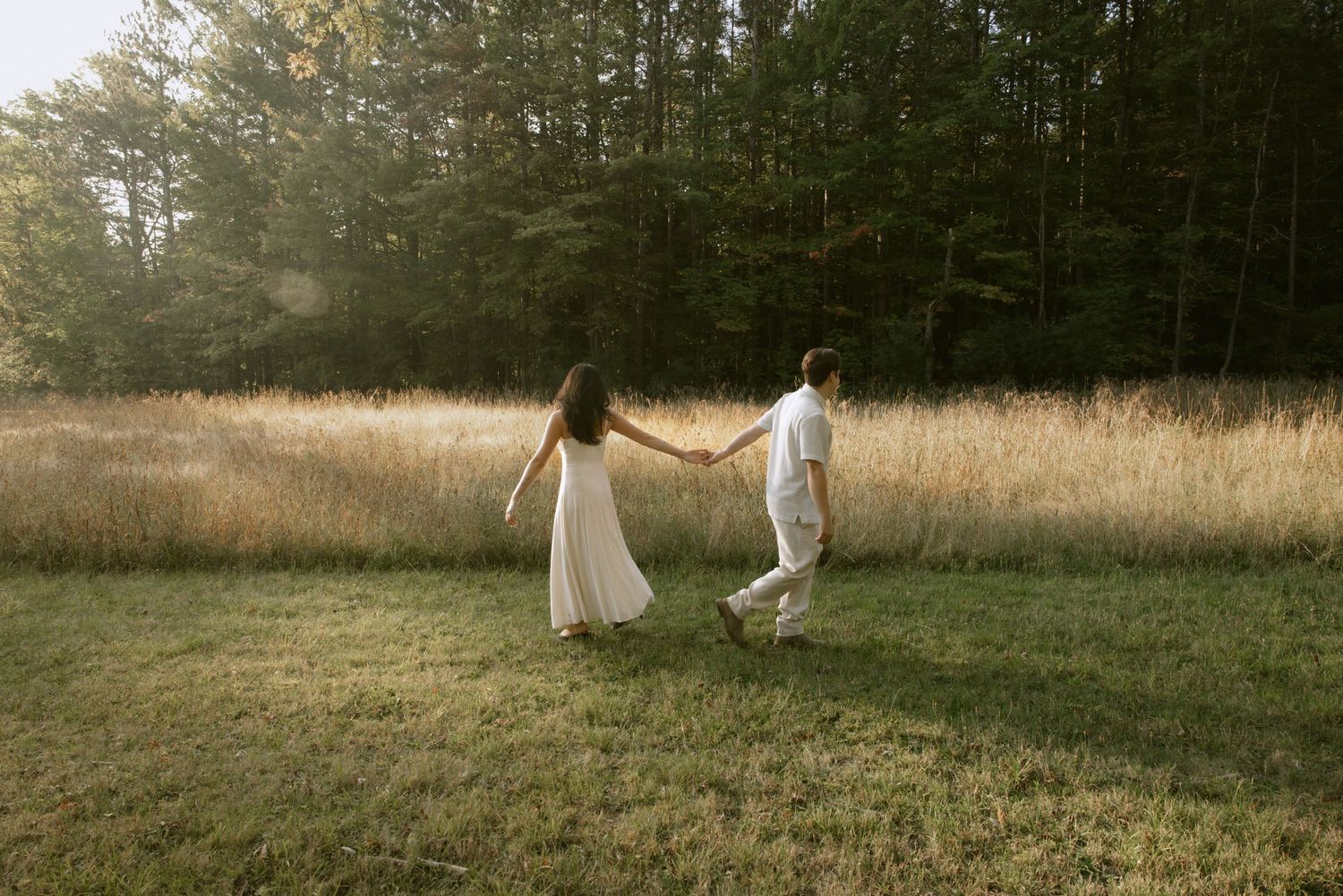 Distant view of two figures in white walking across a sunlit meadow near trees. Rochester NY authentic engagement photography