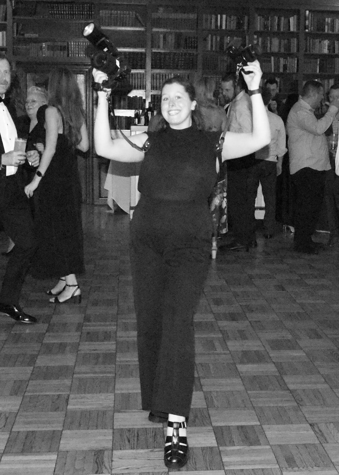 Woman in black dress holds two cameras above her head on a dance floor at an elegant party.