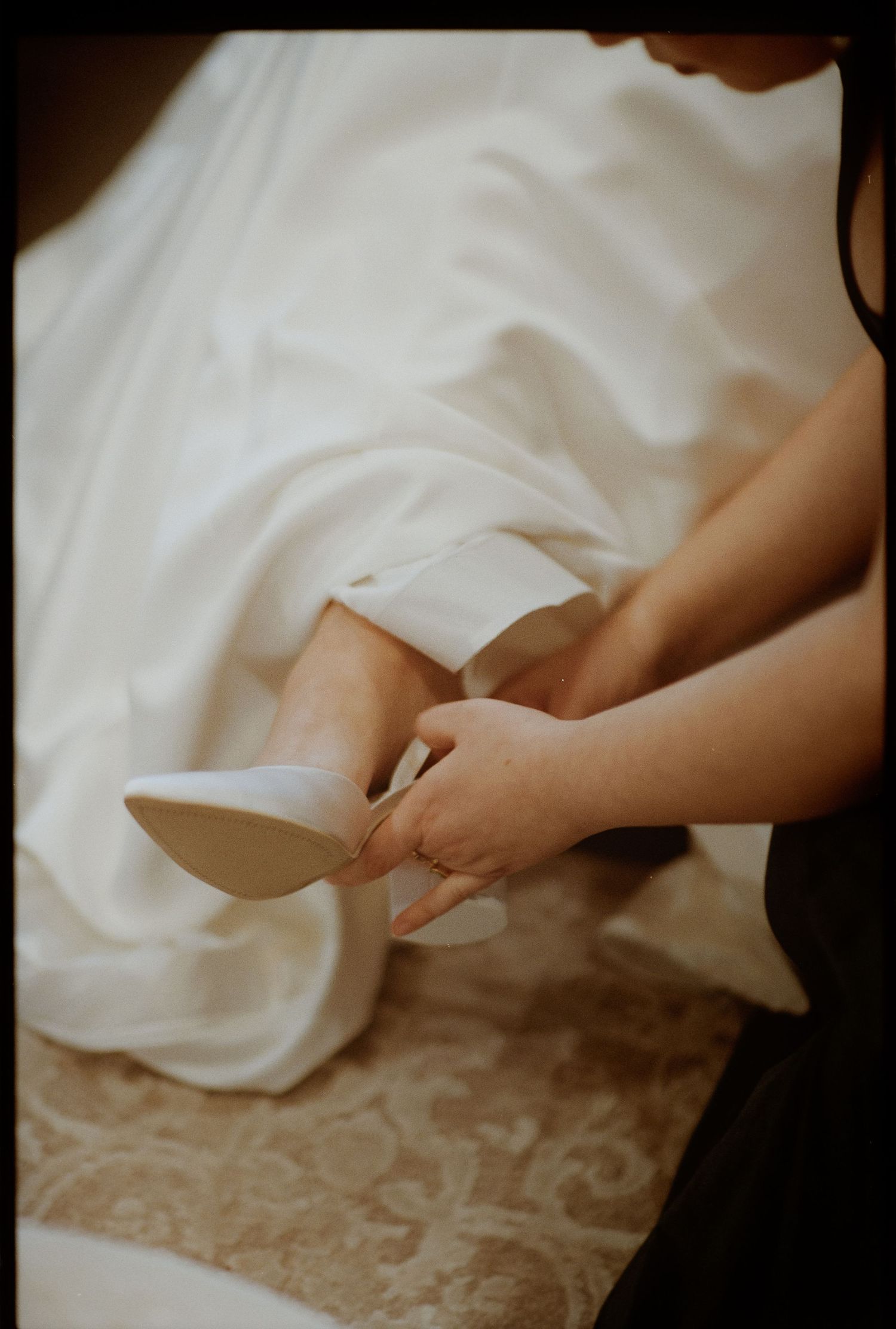 Hands helping to put on a white wedding shoe with flowing dress fabric in the background.