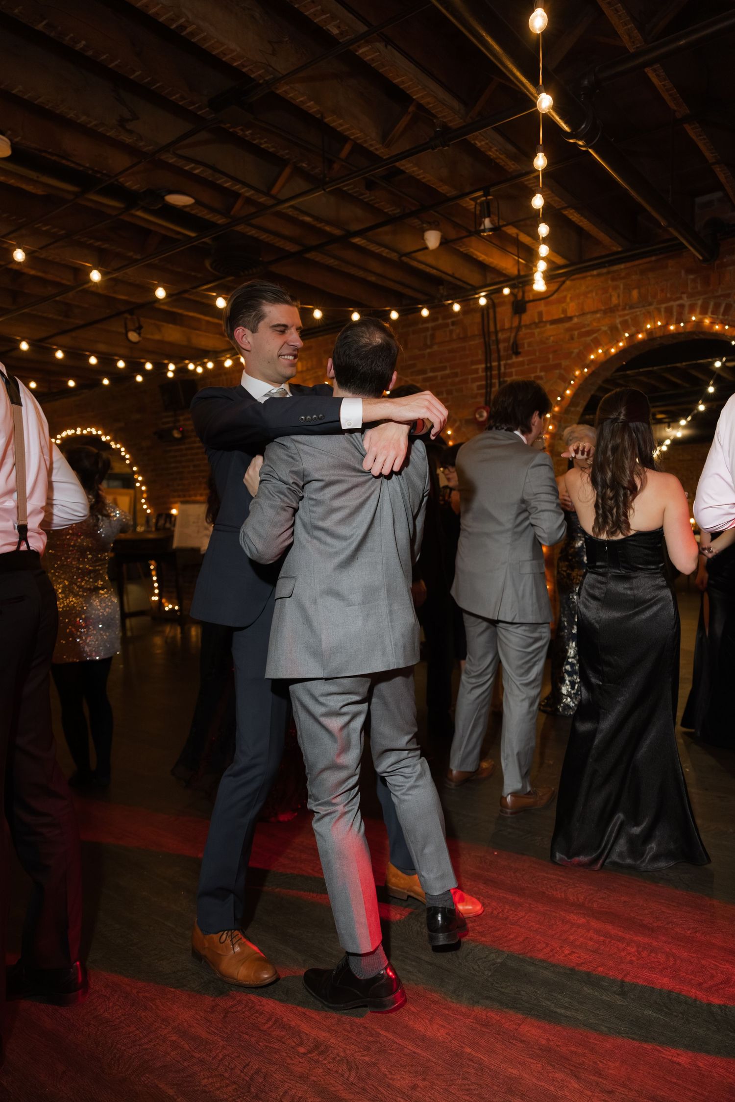 A couple dressed in formal attire dancing under string lights in a rustic indoor setting.