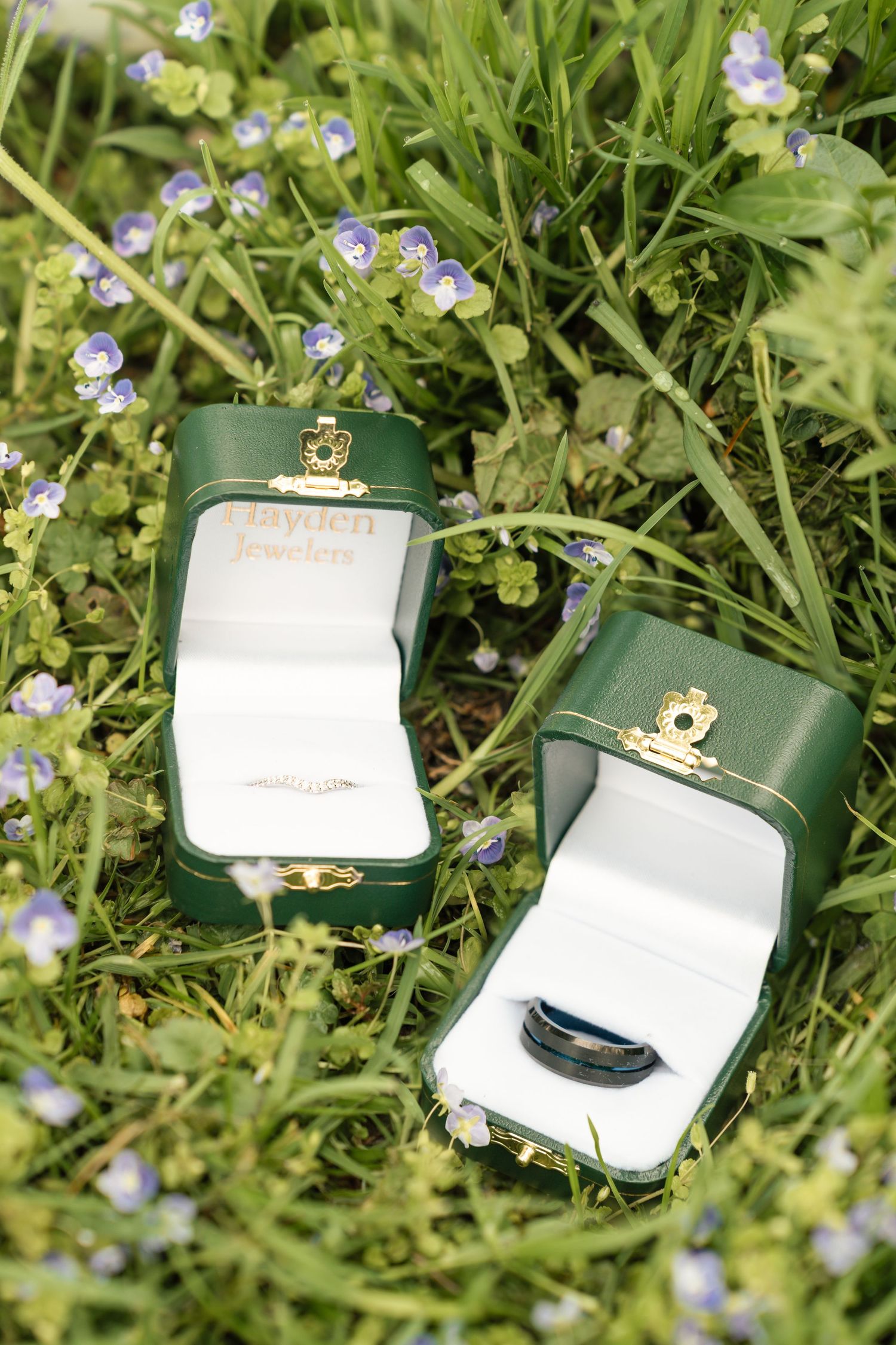Wedding rings displayed in green jewelry boxes among small purple wildflowers in grass. Film wedding photographer NY