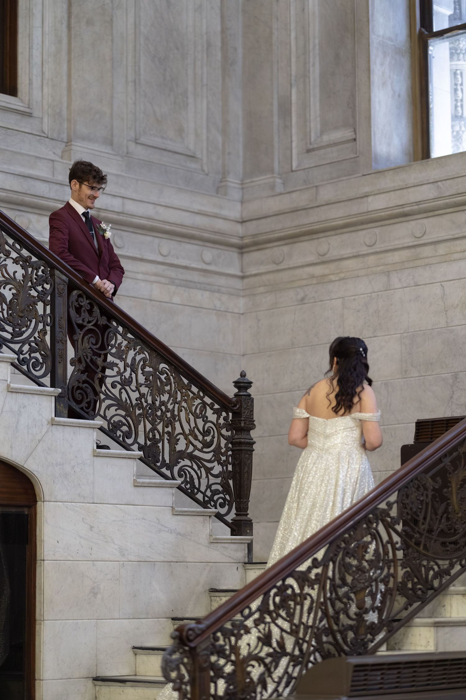 A man in a maroon suit stands on a grand staircase, while a woman in a white dress ascends the stairs.