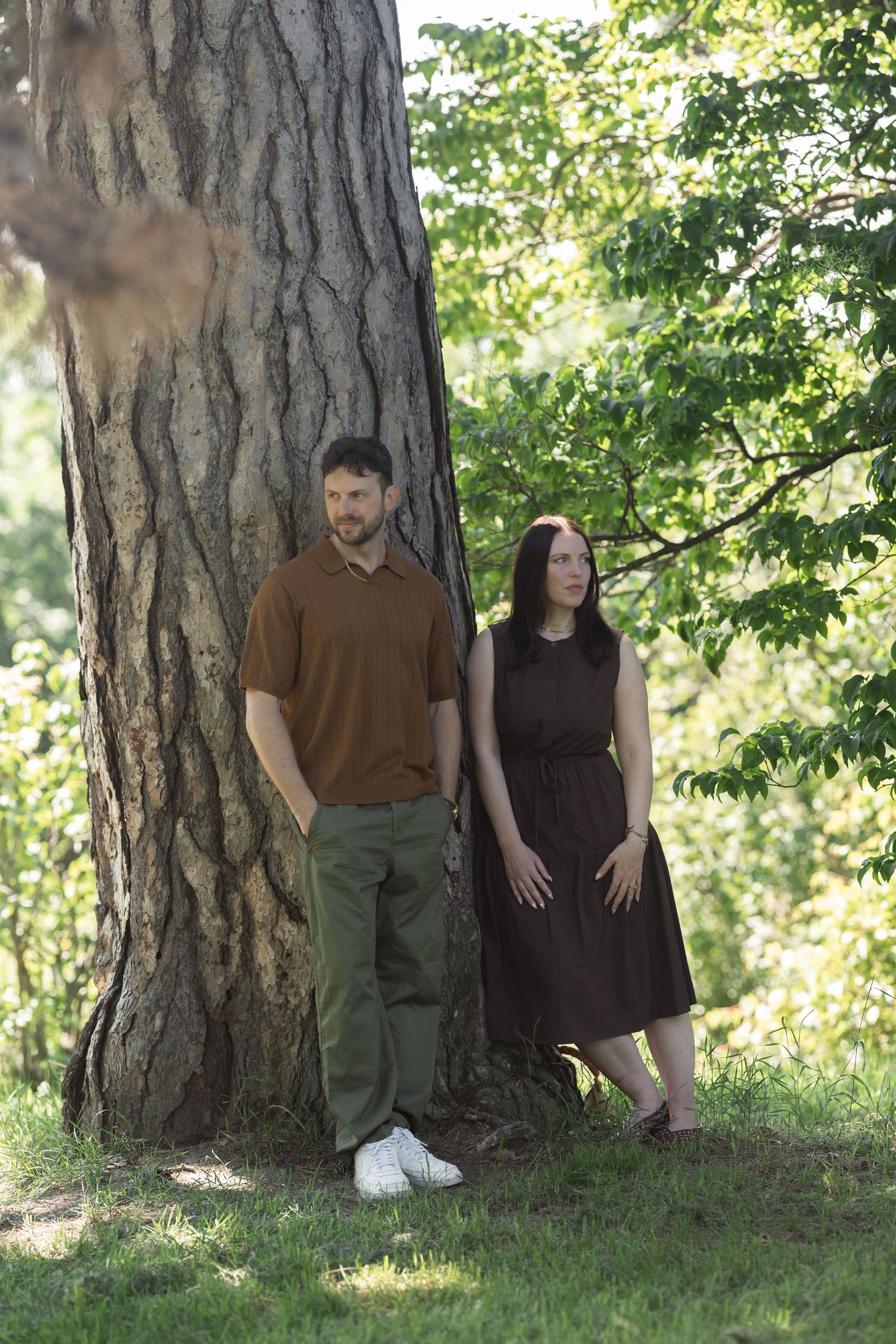 A man and woman stand beside a large tree trunk in a lush green park, he in brown shirt and she in a dark dress.