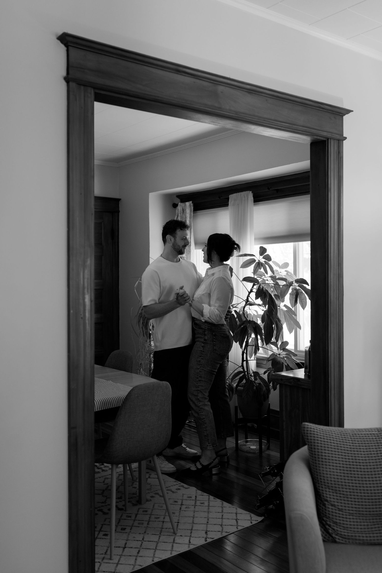 Black and white photo of a couple standing together near a window and houseplant, viewed through a doorway.