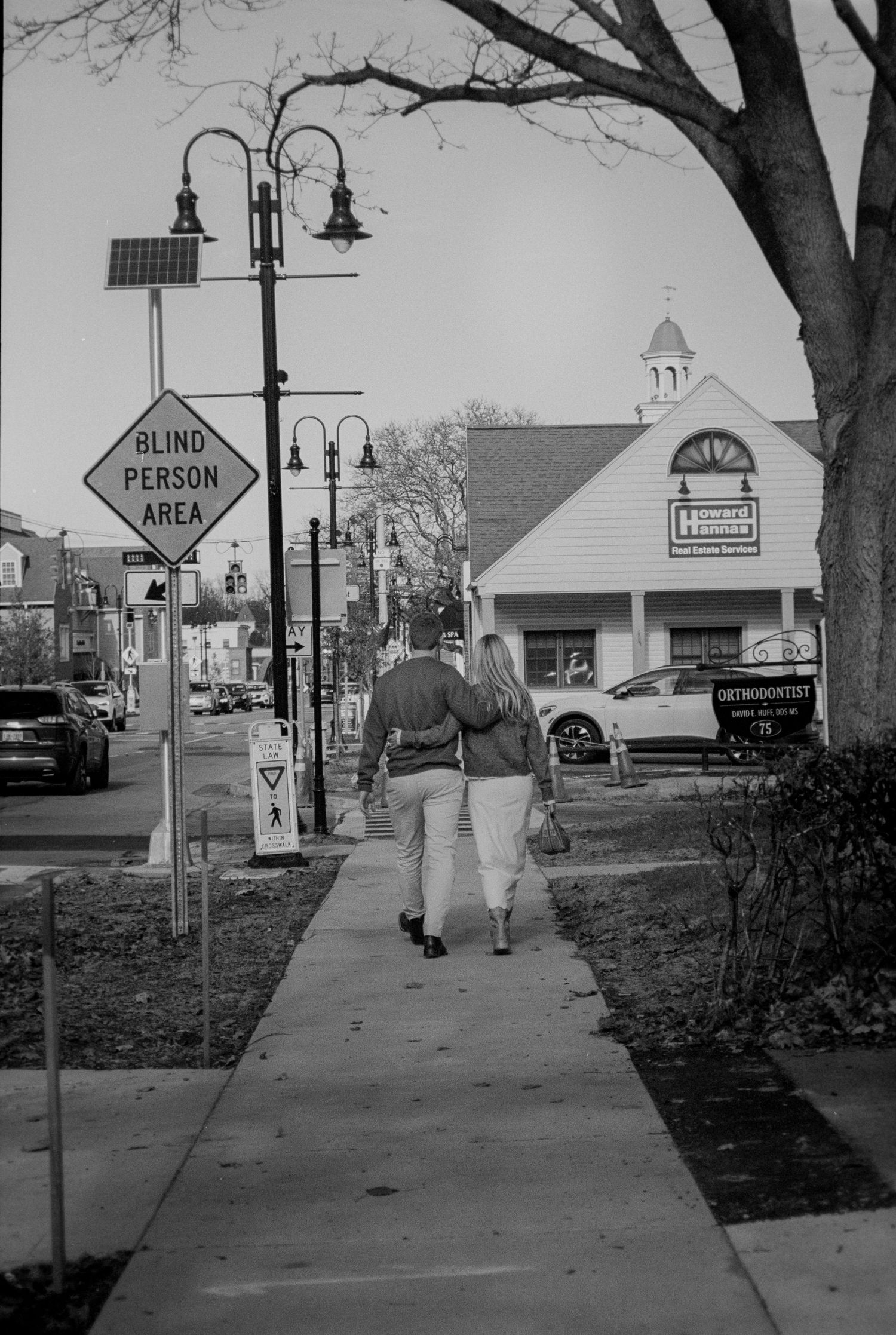 A couple walks hand in hand down a sidewalk past a blind person area sign.
