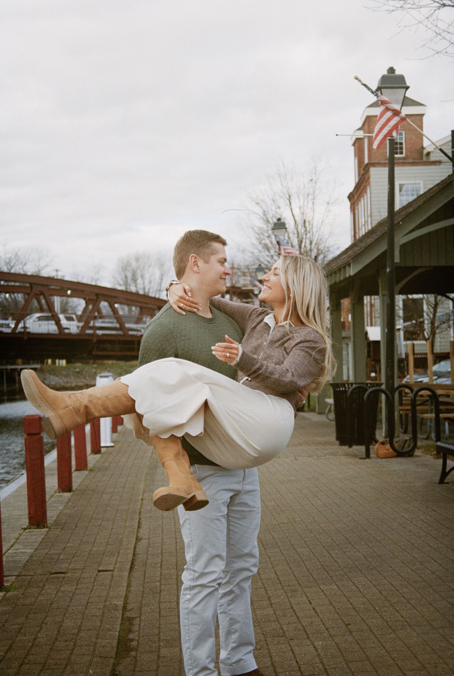 A romantic moment captured by a bridge and lighthouse as a couple embraces in a playful lift pose.