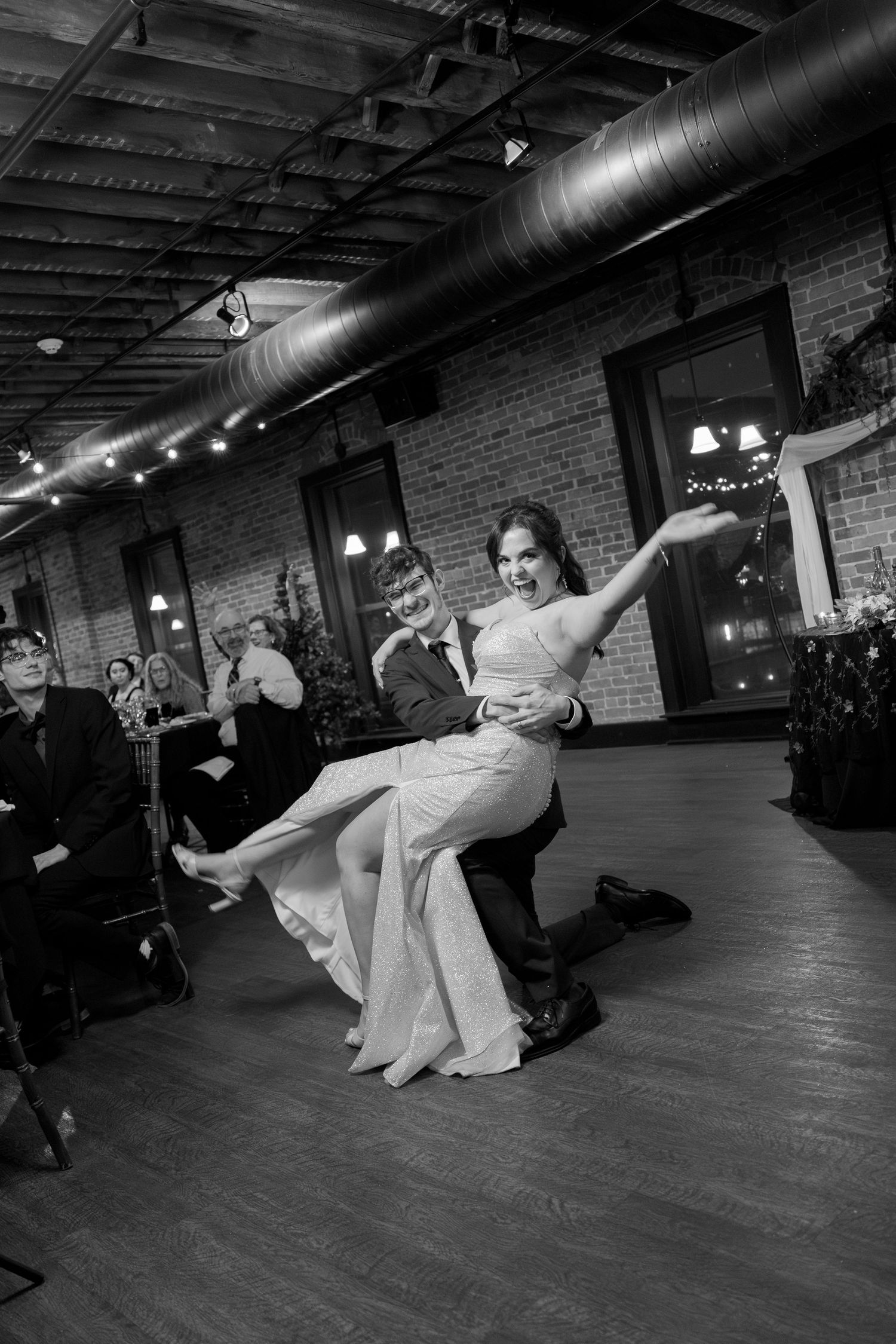 A black and white photograph of a woman in a long dress dancing on a wooden dance floor in a dimly lit room.