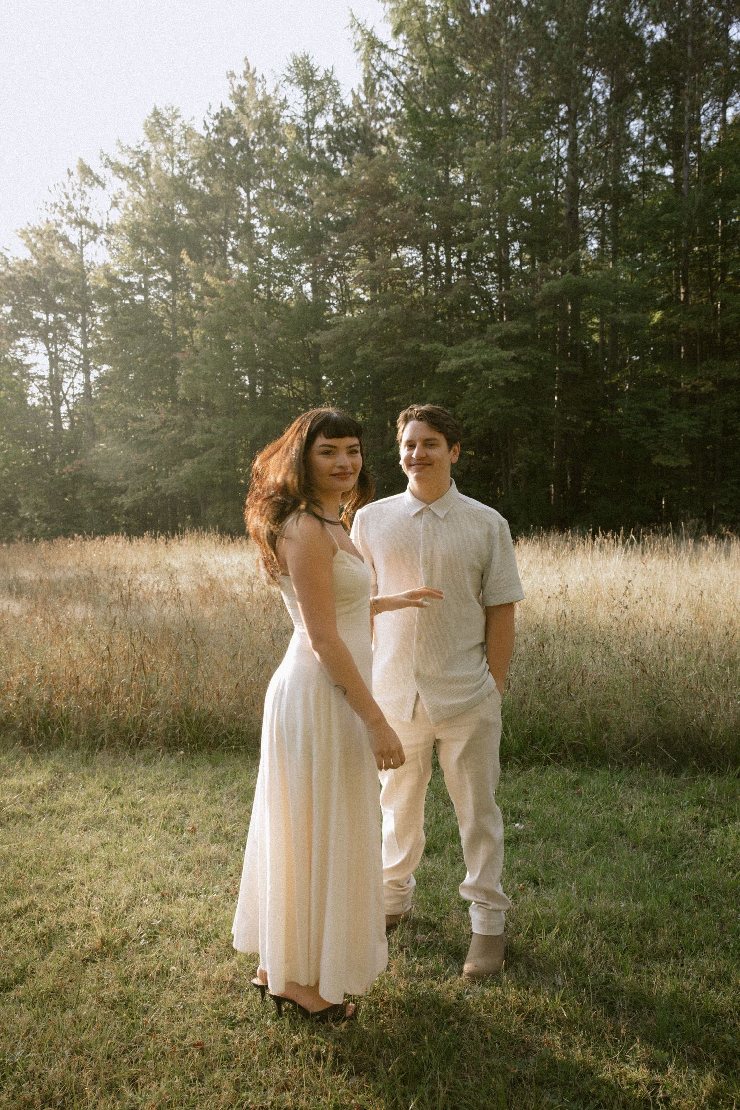 A couple in coordinating white outfits poses together in a grassy field with forest backdrop.  Rochester NY film engagement photography