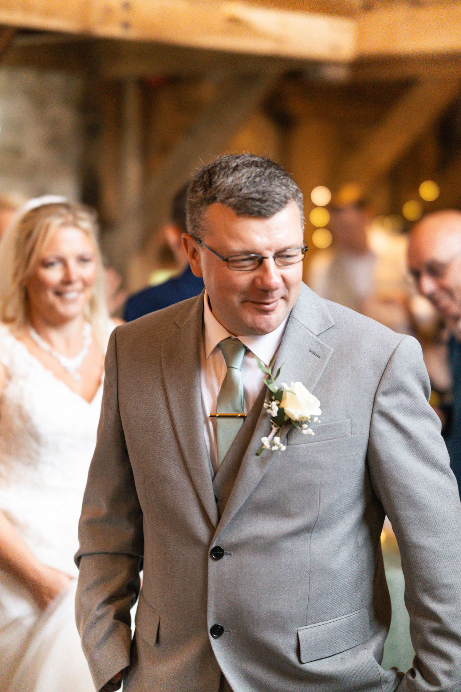 Candid moment of groom walking through the wedding breakfast to the top table with bride looking on from the background