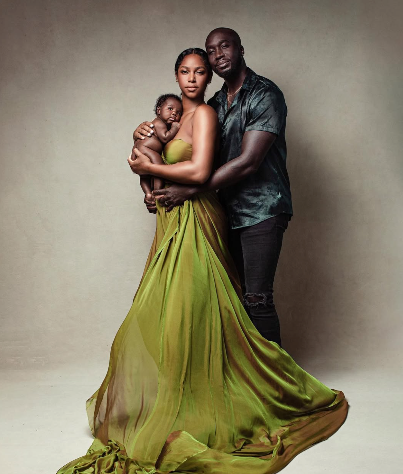 Elegant family portrait of parents in green gown and dark shirt holding newborn baby against neutral background.