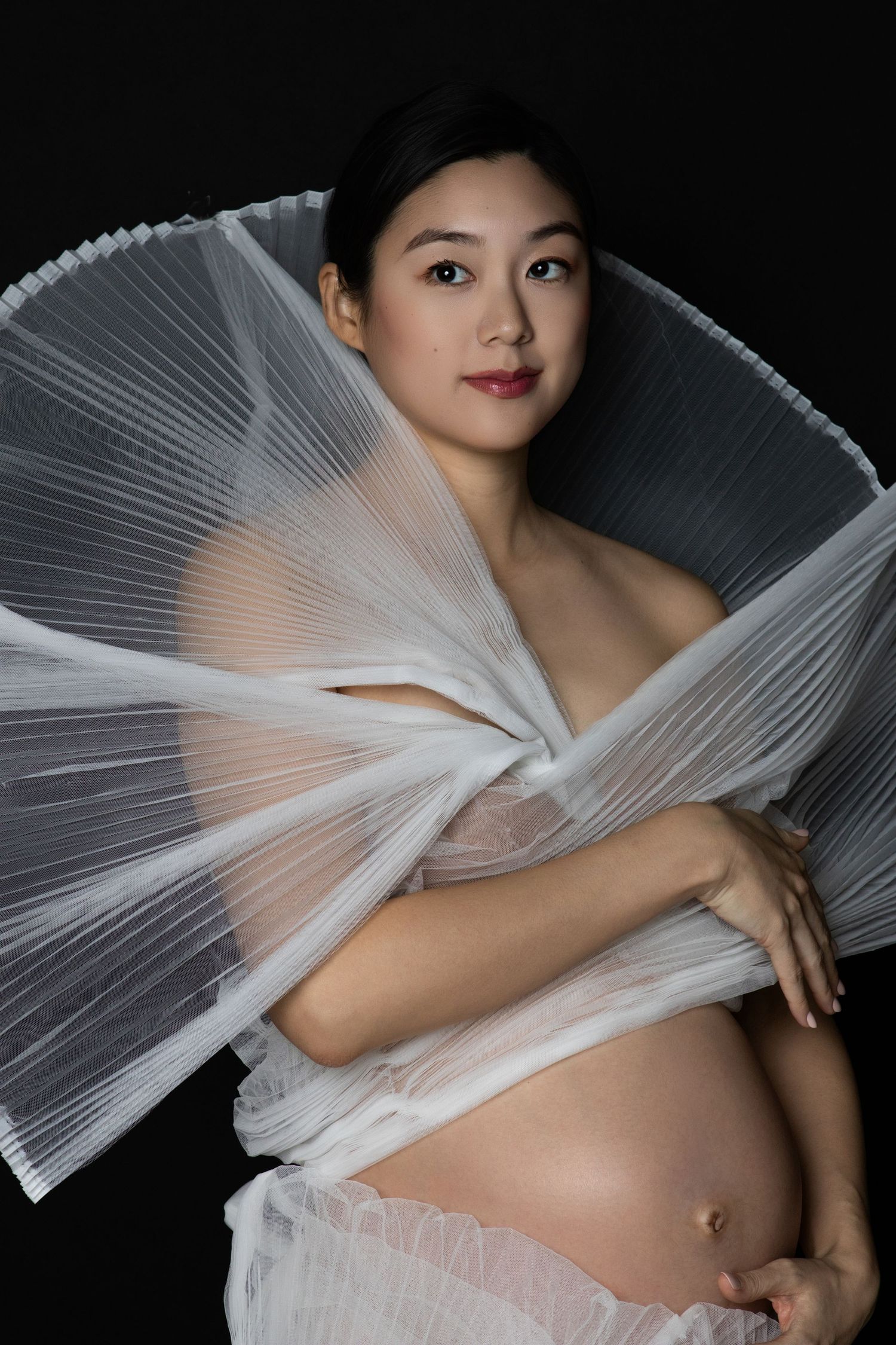 Pregnant Asian woman wearing elegant white pleated fabric wrap poses gracefully against black background.
