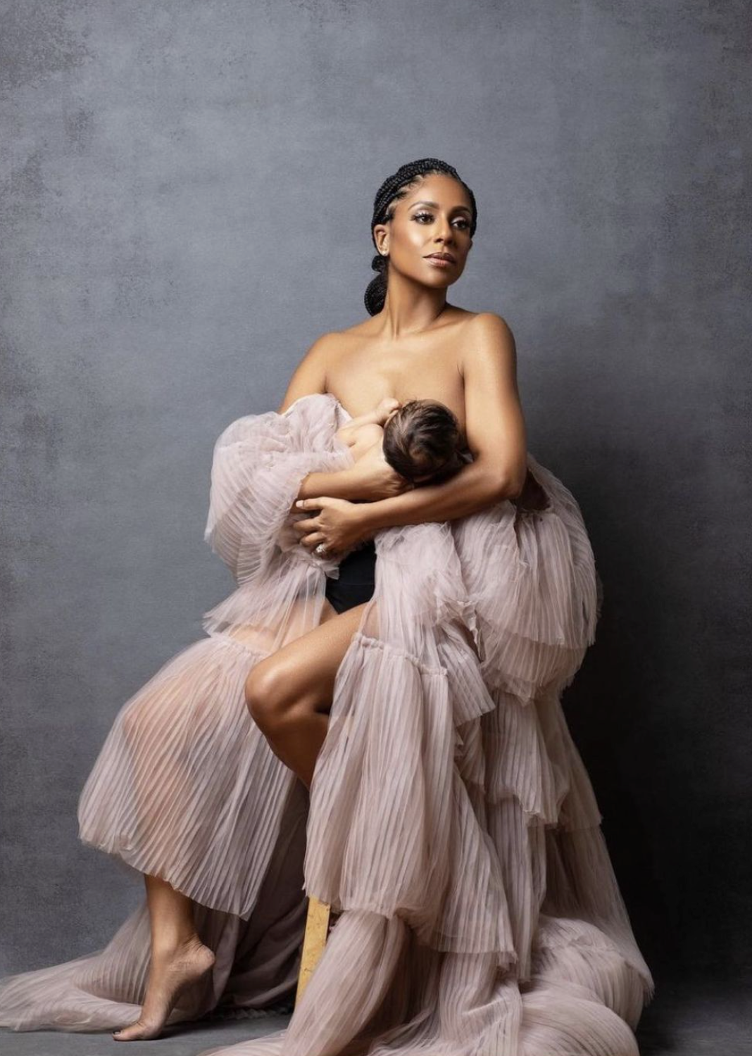 Elegant maternity photo of a woman in a flowing tulle gown breastfeeding her newborn baby in a studio setting.