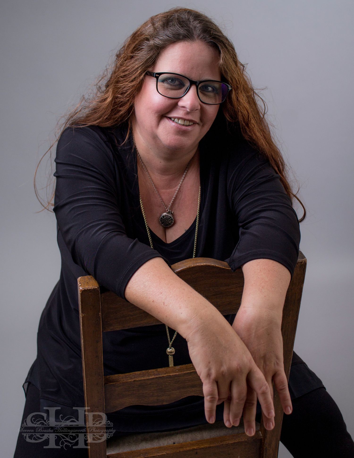 Professional portrait of woman in black attire sitting casually on wooden chair against gray background.