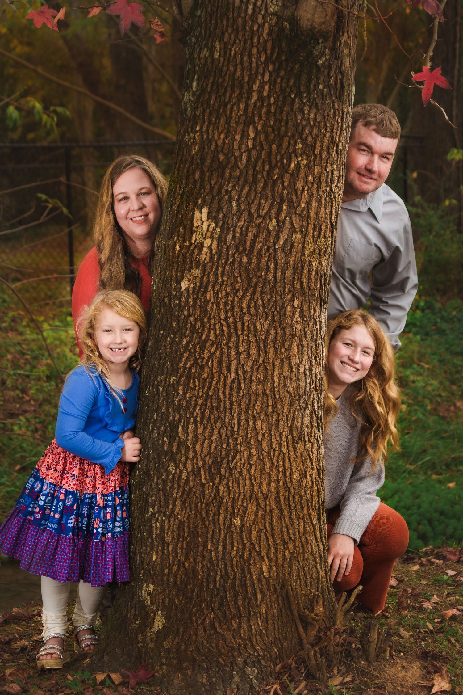 Family poses playfully around large tree trunk in autumn setting with colorful leaves.