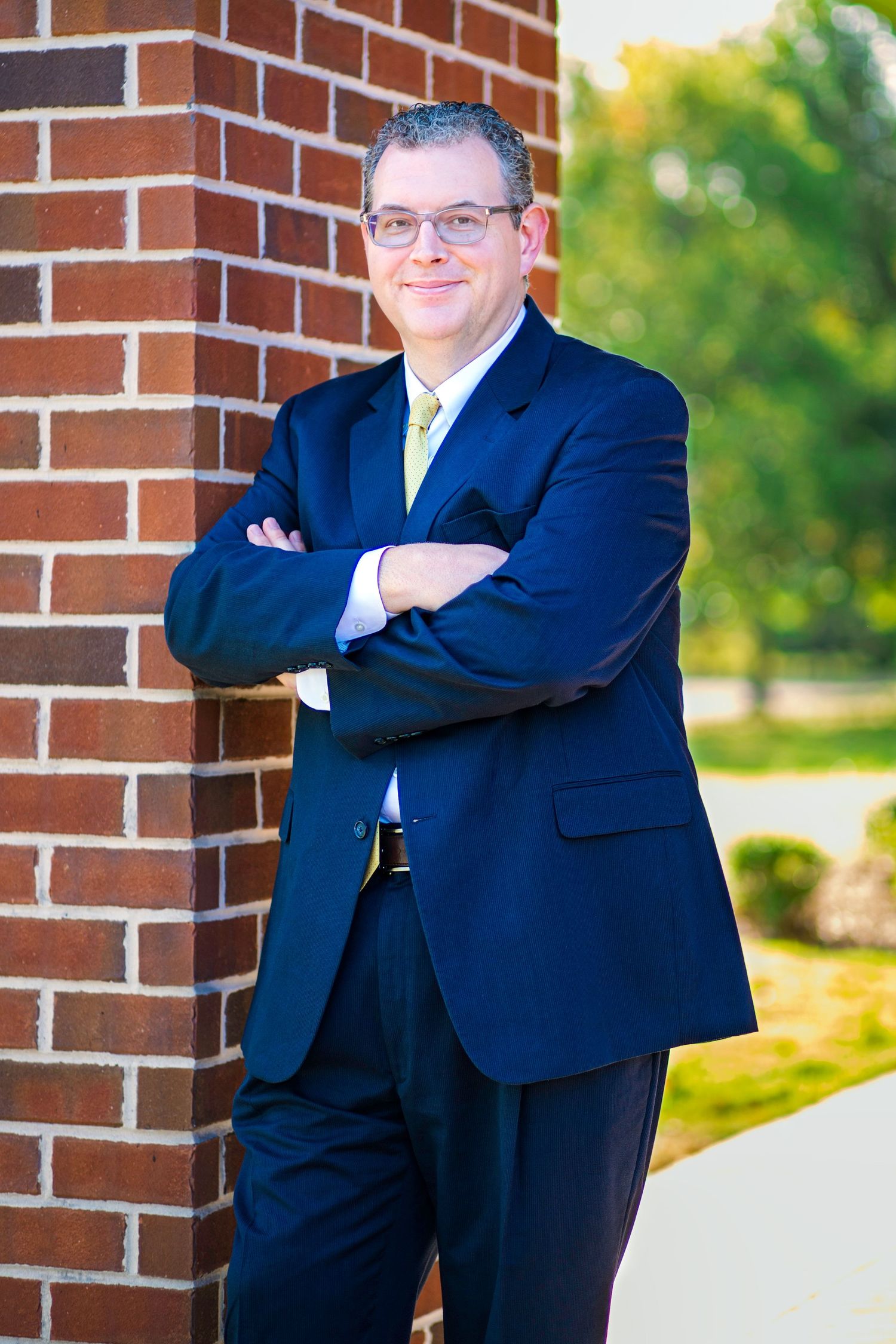 Professional portrait of someone in a navy suit standing against a brick wall.