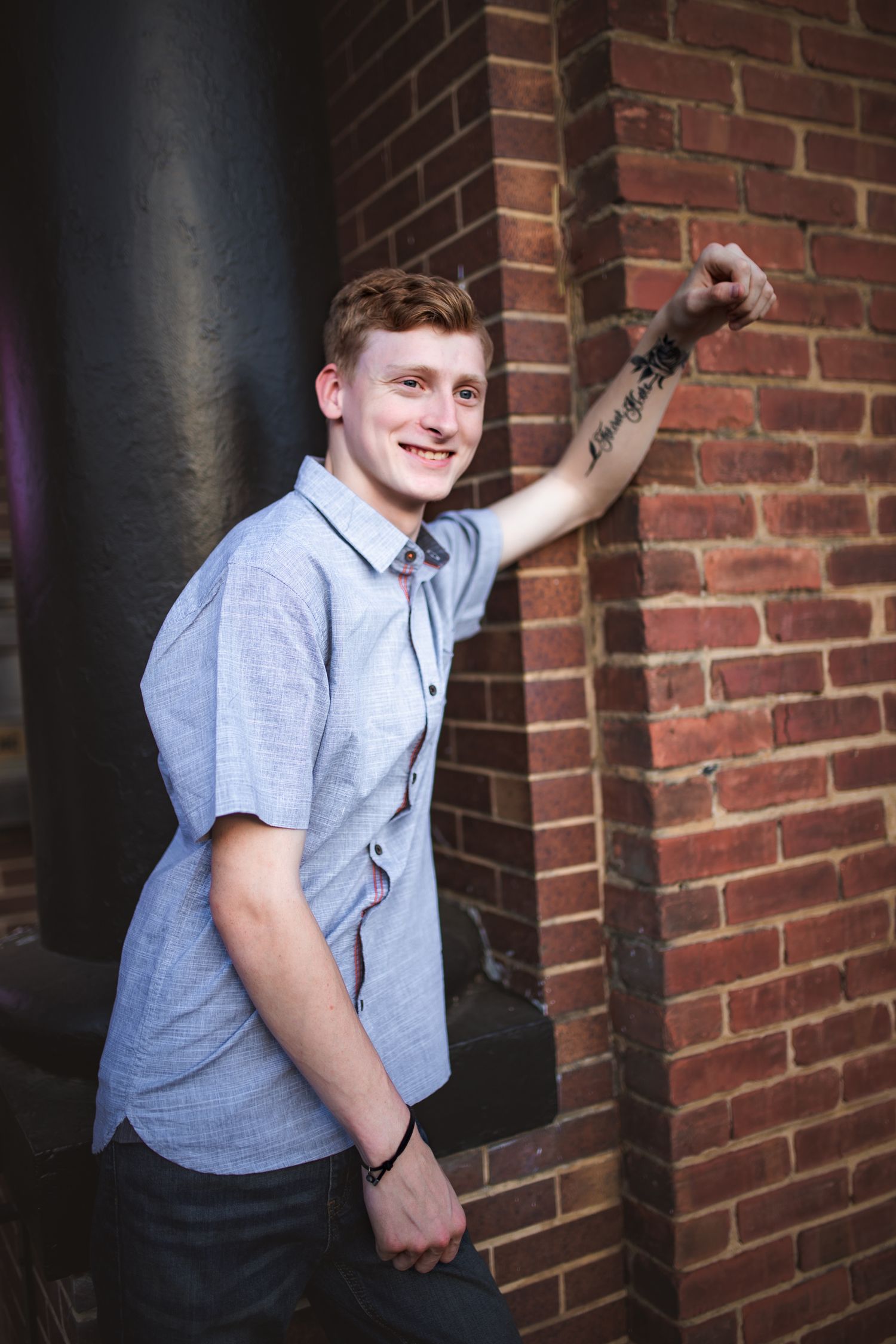 Casual portrait of person in light blue button-down shirt leaning against brick wall with visible arm tattoo.