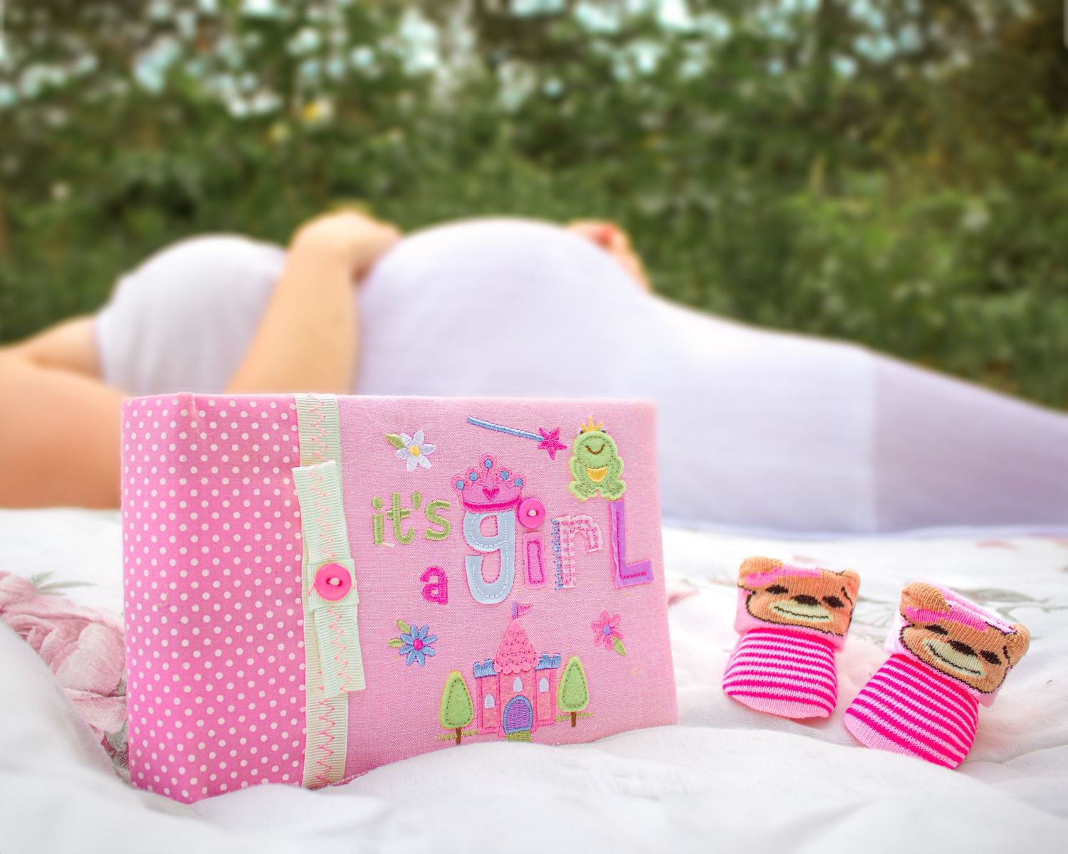 Pink maternity photo with baby shoes and decorative first birthday card on white blanket.