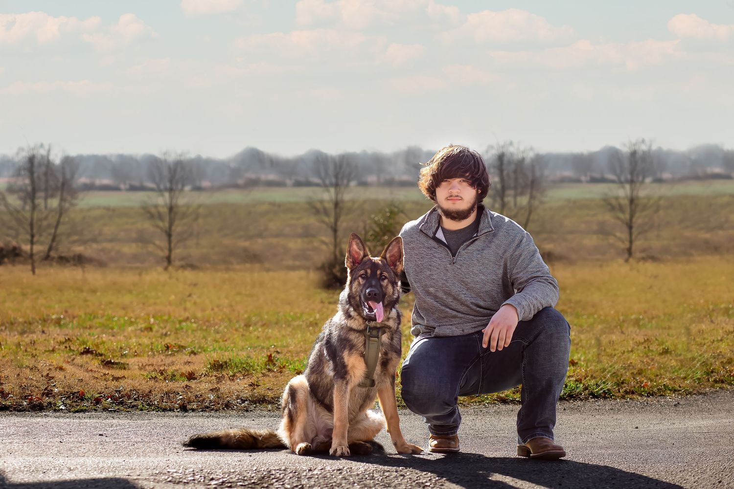A person wearing a gray sweater kneels beside a German Shepherd dog on a path with meadows in the background.
