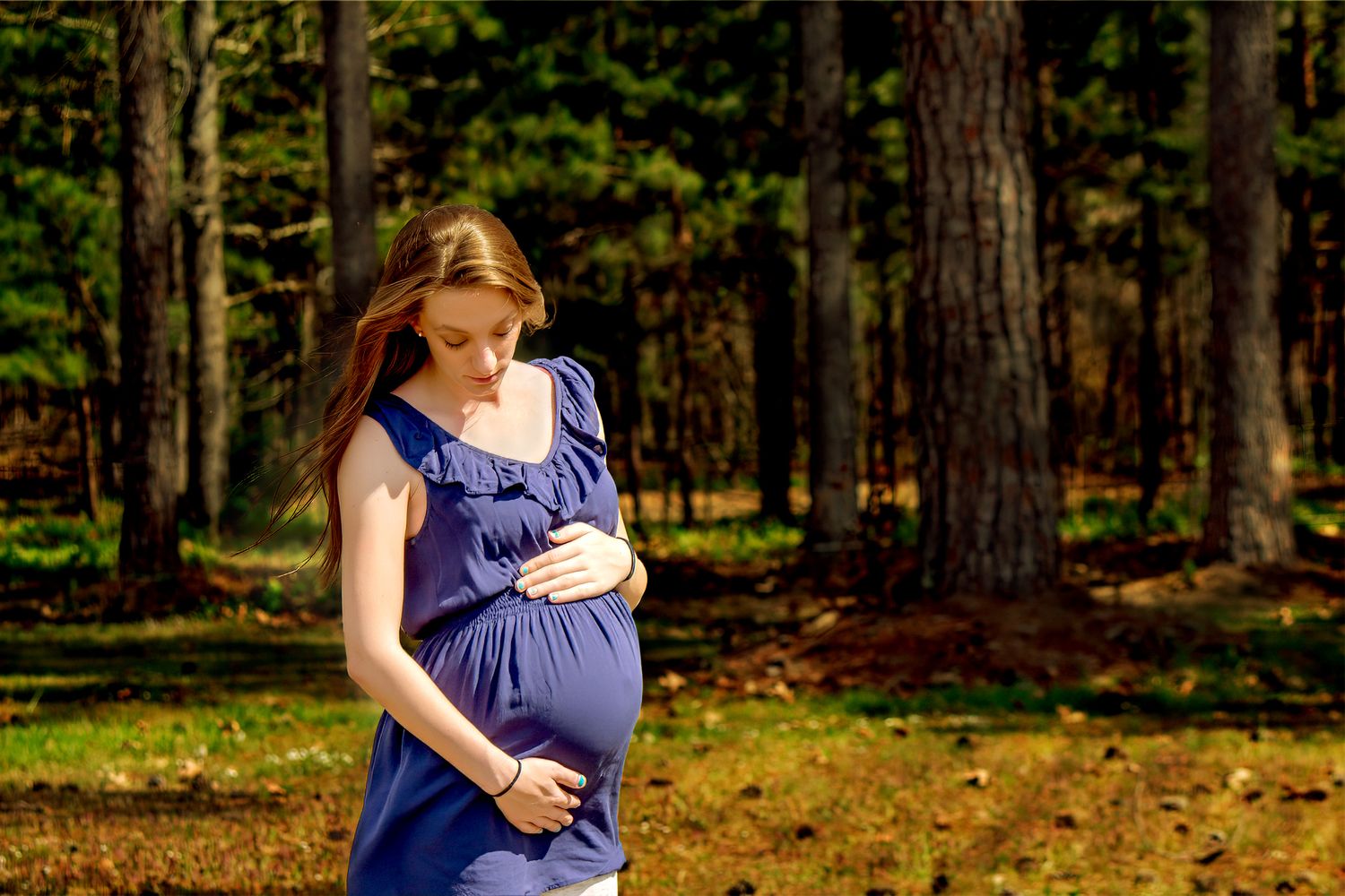 Woman in navy blue dress poses in sunlit forest during maternity photoshoot.