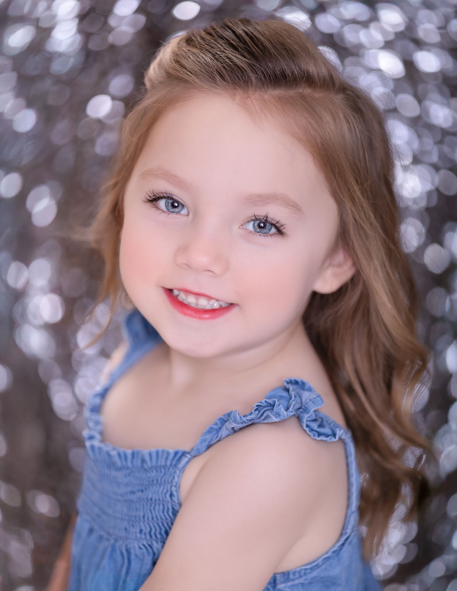 Child in a ruffled blue dress poses for portrait against sparkly silver bokeh background.