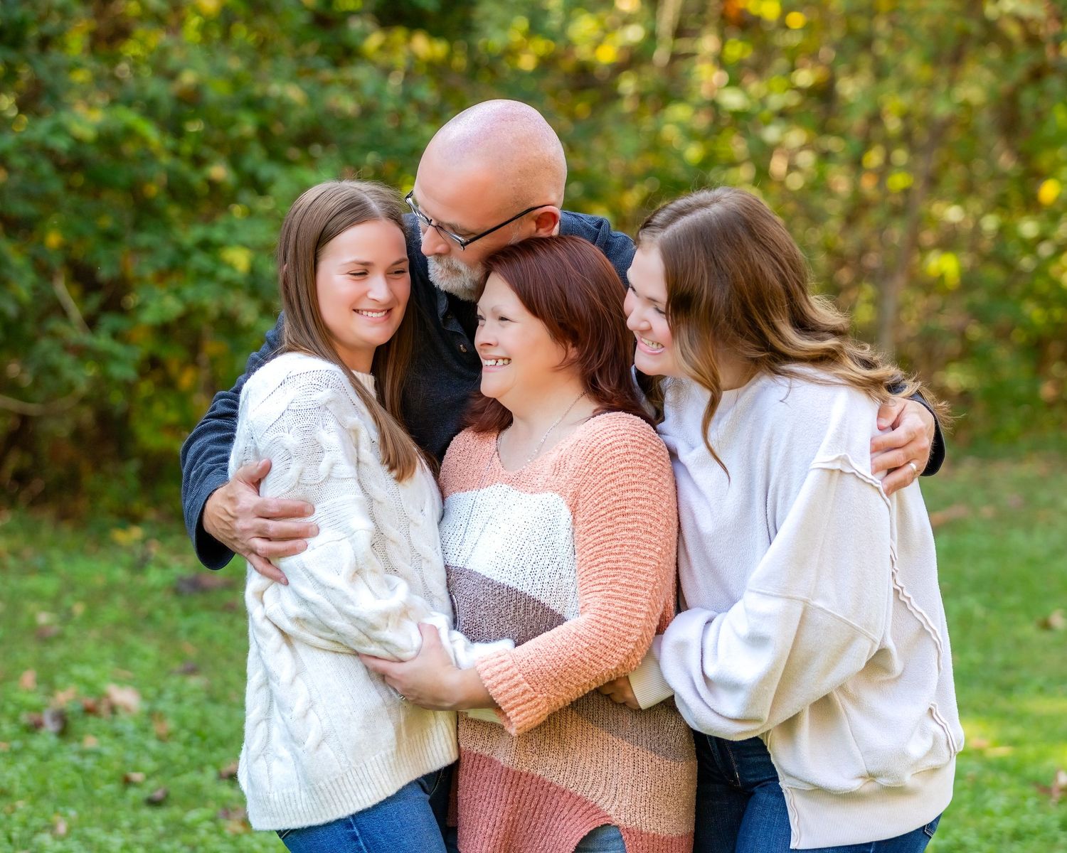Happy family of four hugging outdoors in autumn, parents with two teenage daughters smiling in a green park setting.