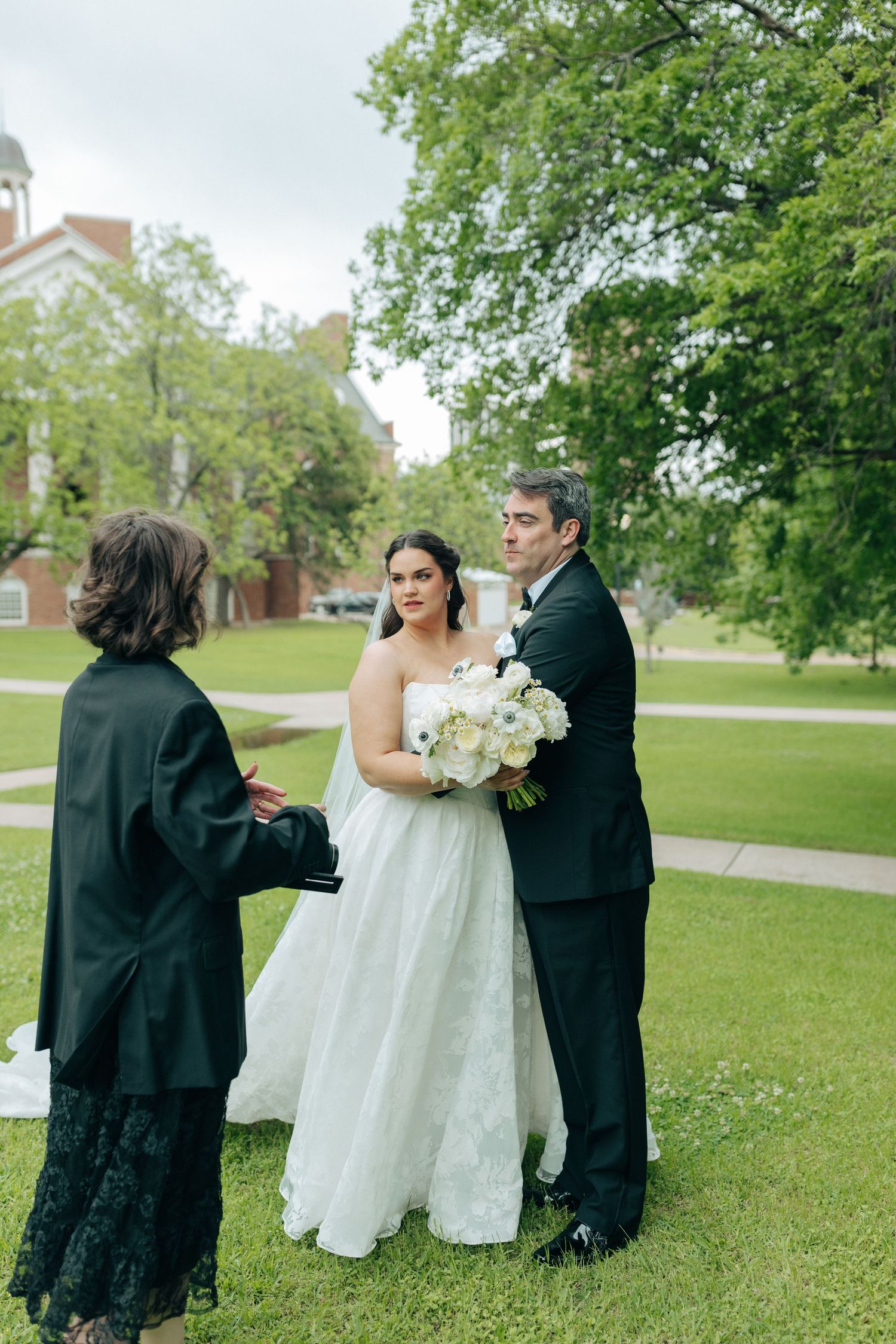 Bride and groom stand together outdoors during intimate wedding ceremony with officiant, surrounded by lush green trees.