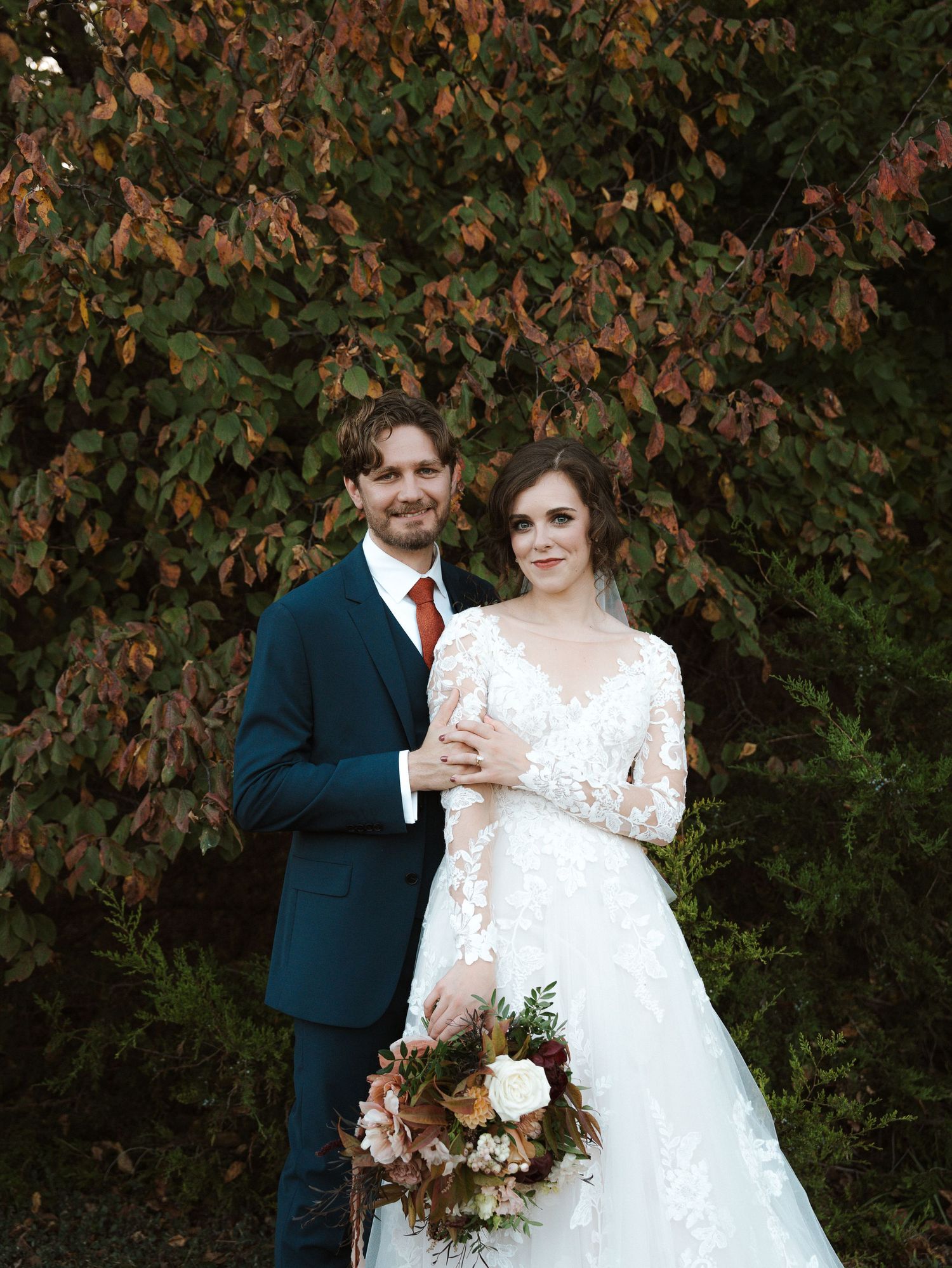 A wedding couple poses in front of autumn foliage with the bride holding a rustic bouquet.