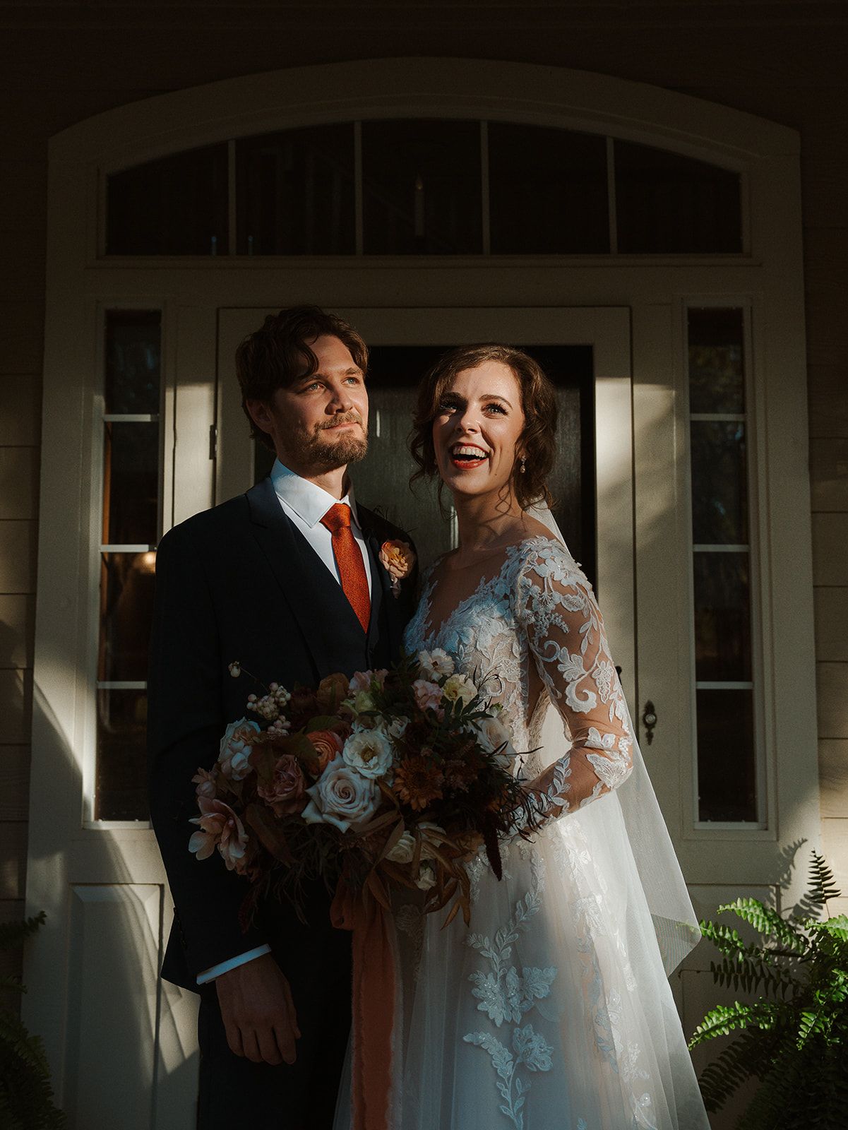 A bride and groom pose in dramatic lighting outside elegant white French doors, she holding a lush bouquet of peach and white blooms.
