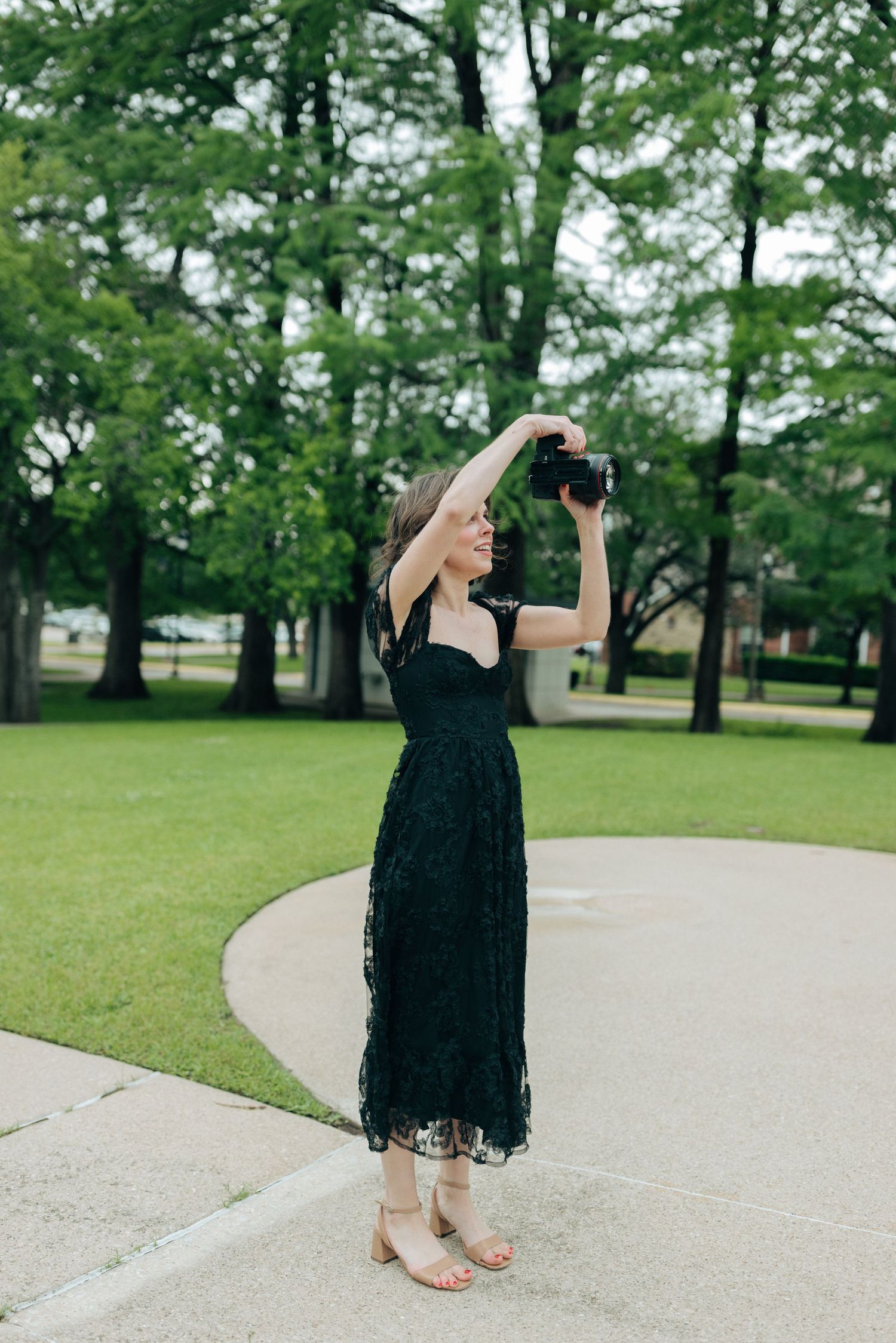Woman in black lace midi dress photographing outdoors in a lush green park, holding camera above her head.