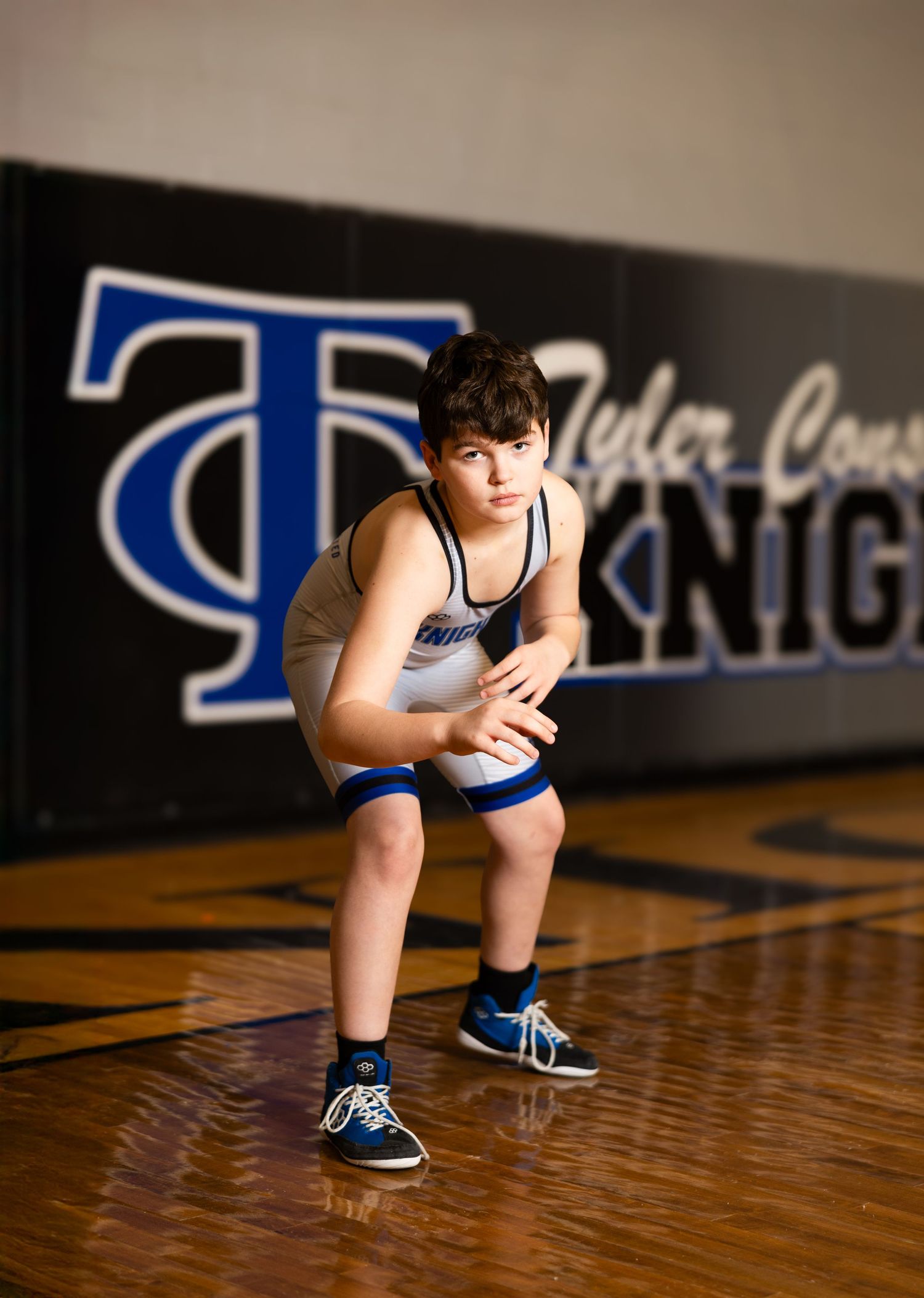 A wrestler in blue uniform crouches in ready position on a gymnasium floor with team logo visible.